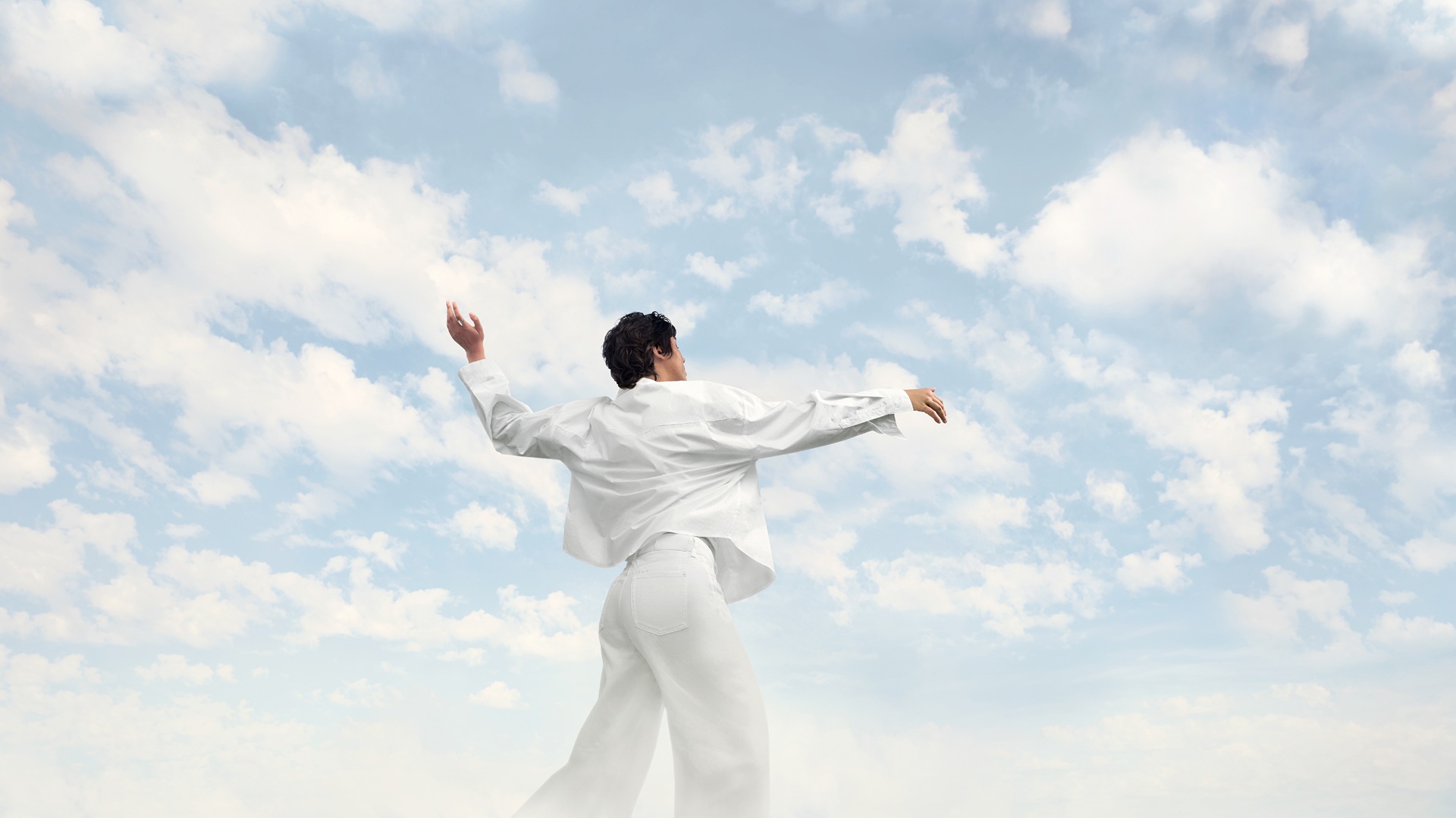 Pessoa vestida de branco dançando ao ar livre sob um céu azul com nuvens claras, em gesto expansivo e fluido