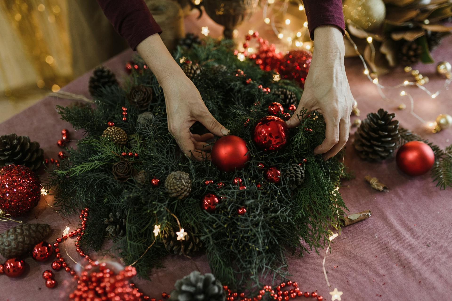 Pessoa montando uma guirlanda de Natal com galhos verdes, pinhas e bolas vermelhas sobre uma mesa decorada com luzes e enfeite