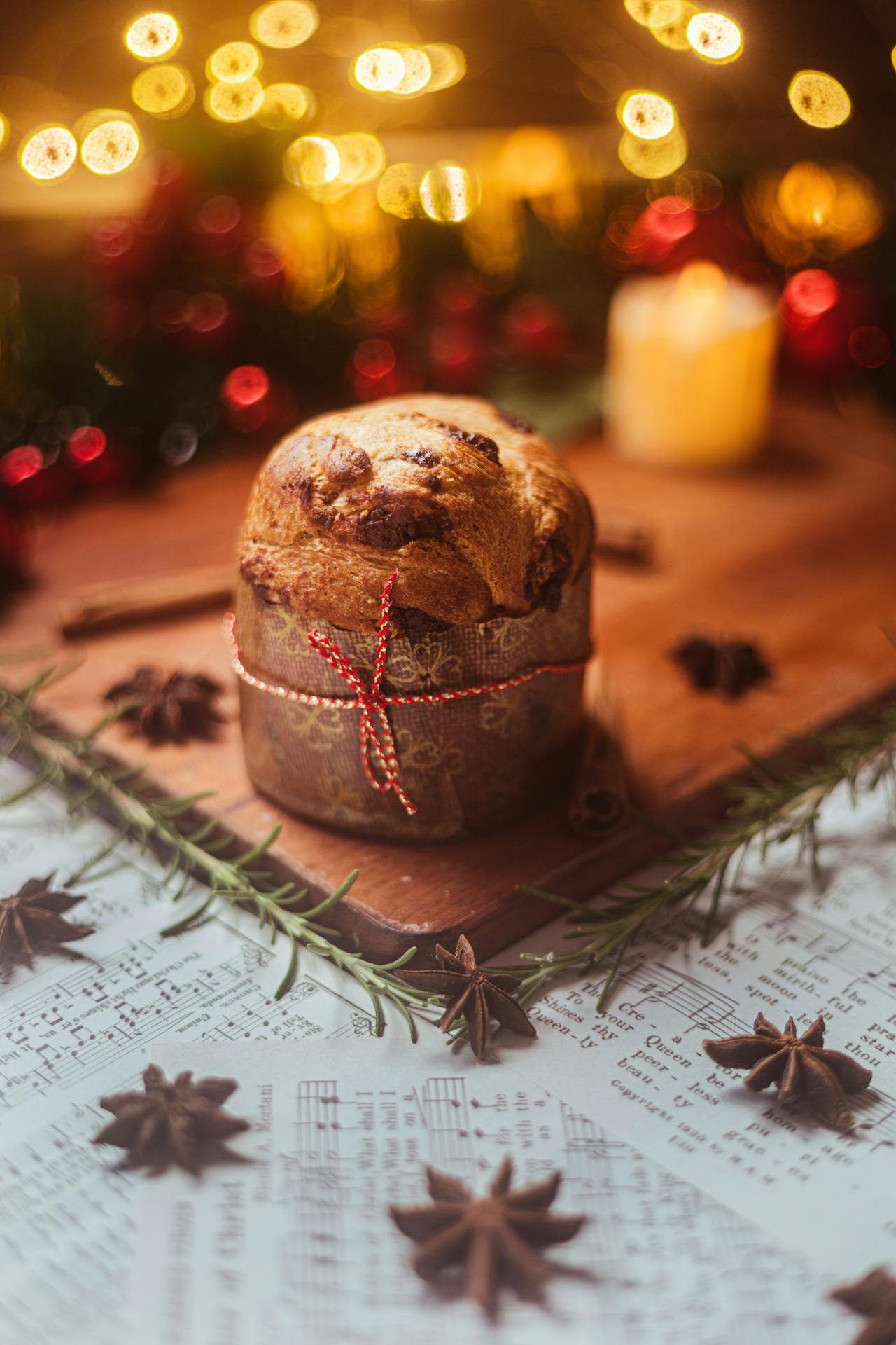 Panetone com pedaços de chocolate, amarrado com cordão vermelho, sobre tábua de madeira decorada com alecrim, anis-estrelado e partituras musicais. Ao fundo, luzes douradas e uma vela acesa criam um clima natalino aconchegante