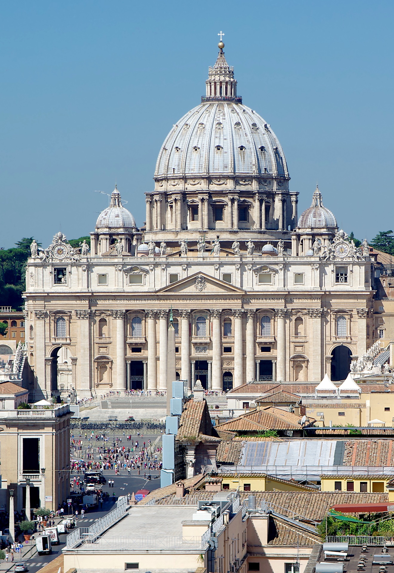 Basílica de São Pedro, em Roma, vista de frente, com a sua grande cúpula central ladeada por torres menores, fachada ornamentada por colunas e esculturas, e movimentação de pessoas na praça ao redor