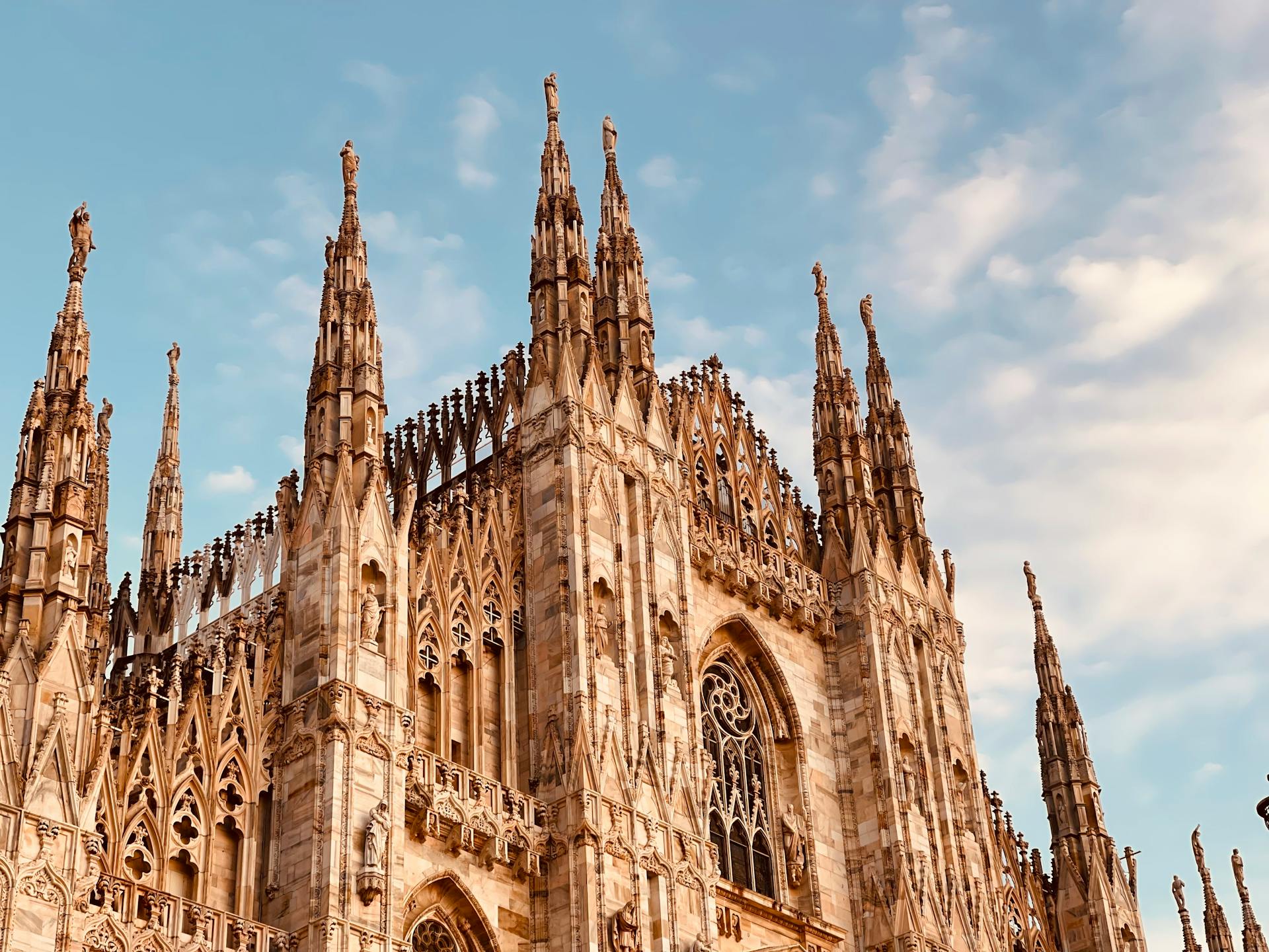 Fotografia em perspectiva da Catedral de Milão (Duomo di Milano), capturada ao pôr do sol, destacando torres e pináculos góticos ricamente ornamentados