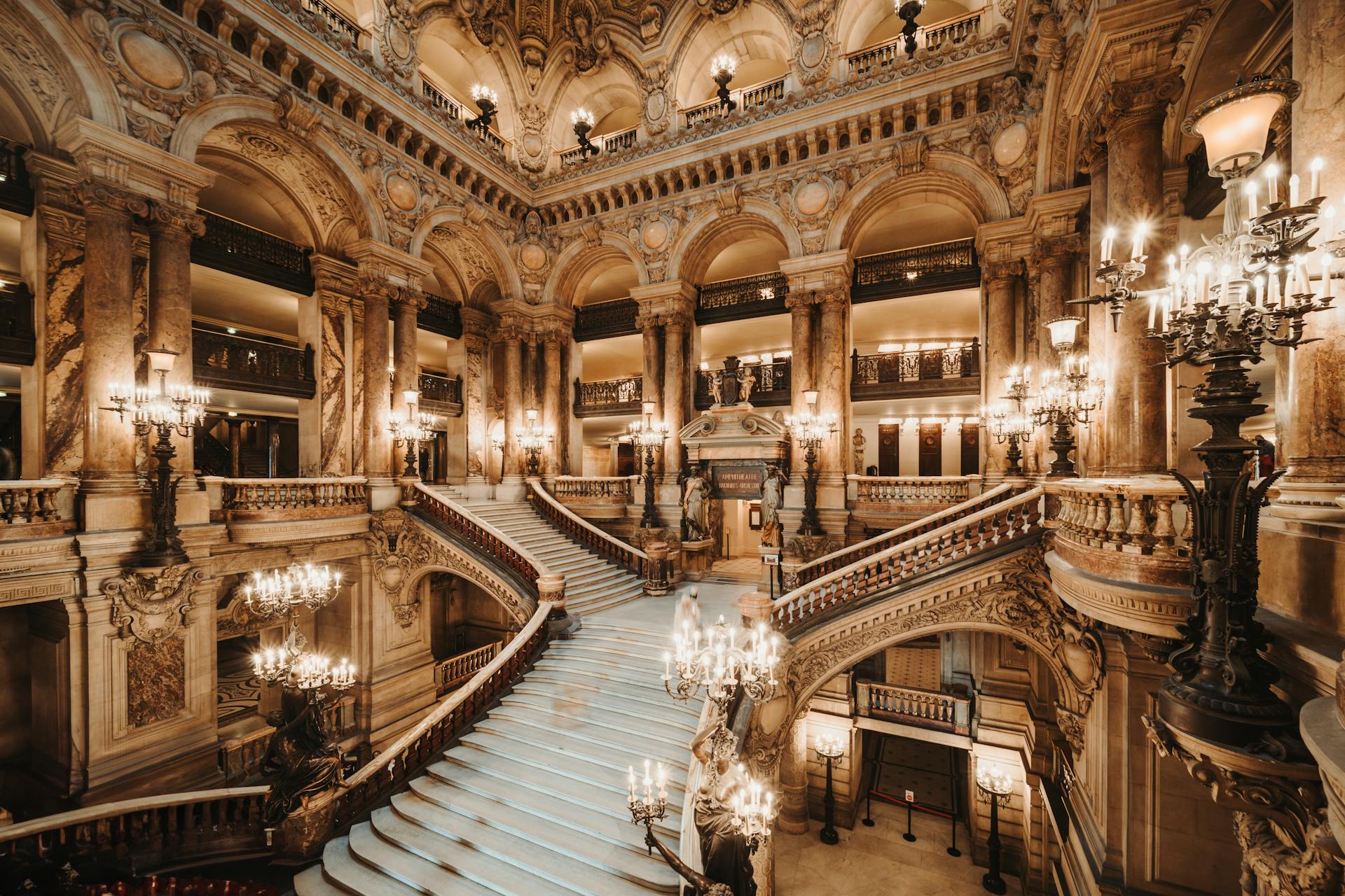 opera garnier Vista interna da Ópera Garnier, em Paris, mostrando a grande escadaria central em mármore branco cercada por colunas ornamentadas, balcões dourados e luminárias em ferro forjado