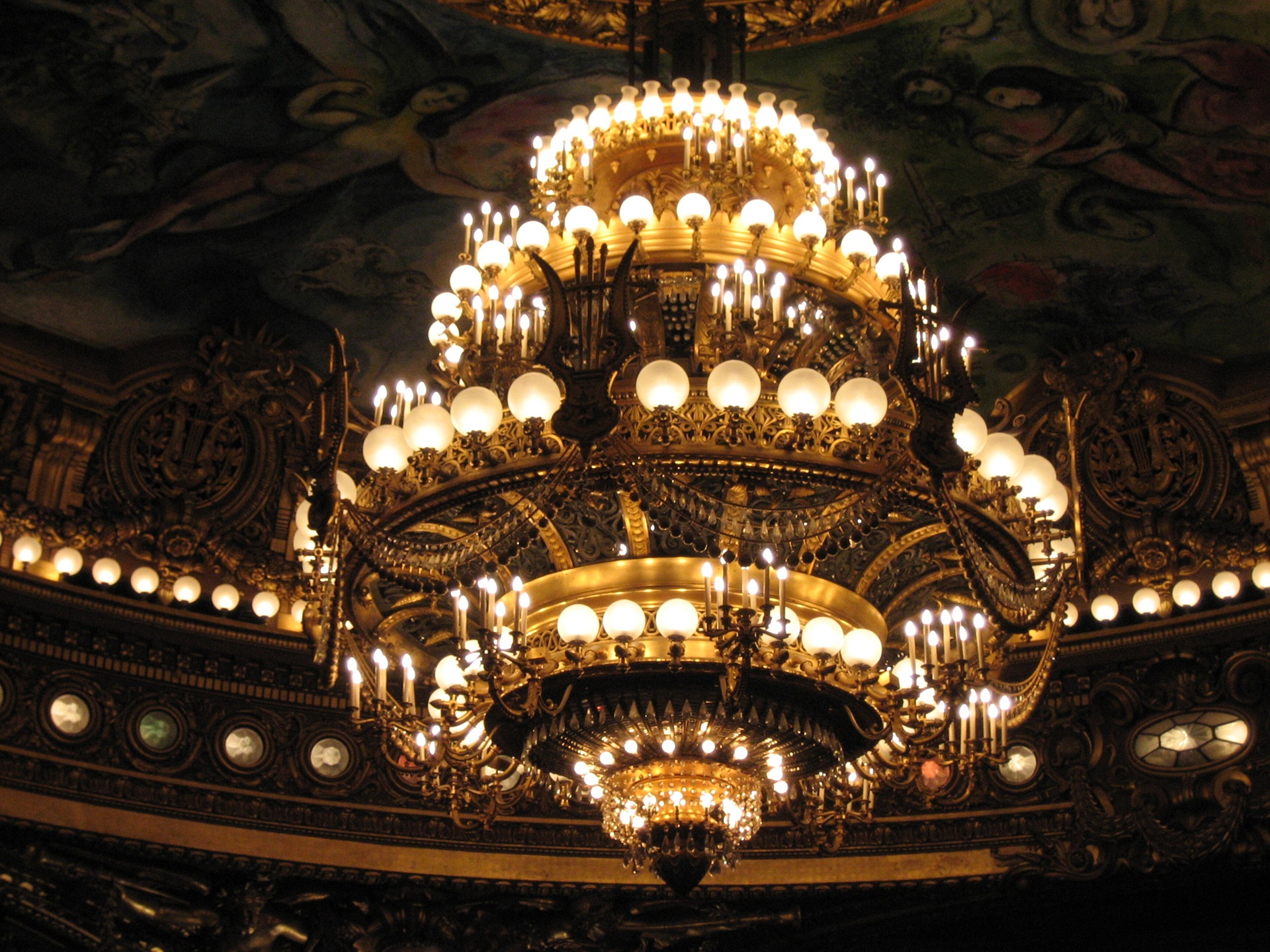 opera garnier Close do imponente lustre central da Ópera Garnier, feito em bronze e cristal, com centenas de lâmpadas acesas e detalhes ornamentais dourados