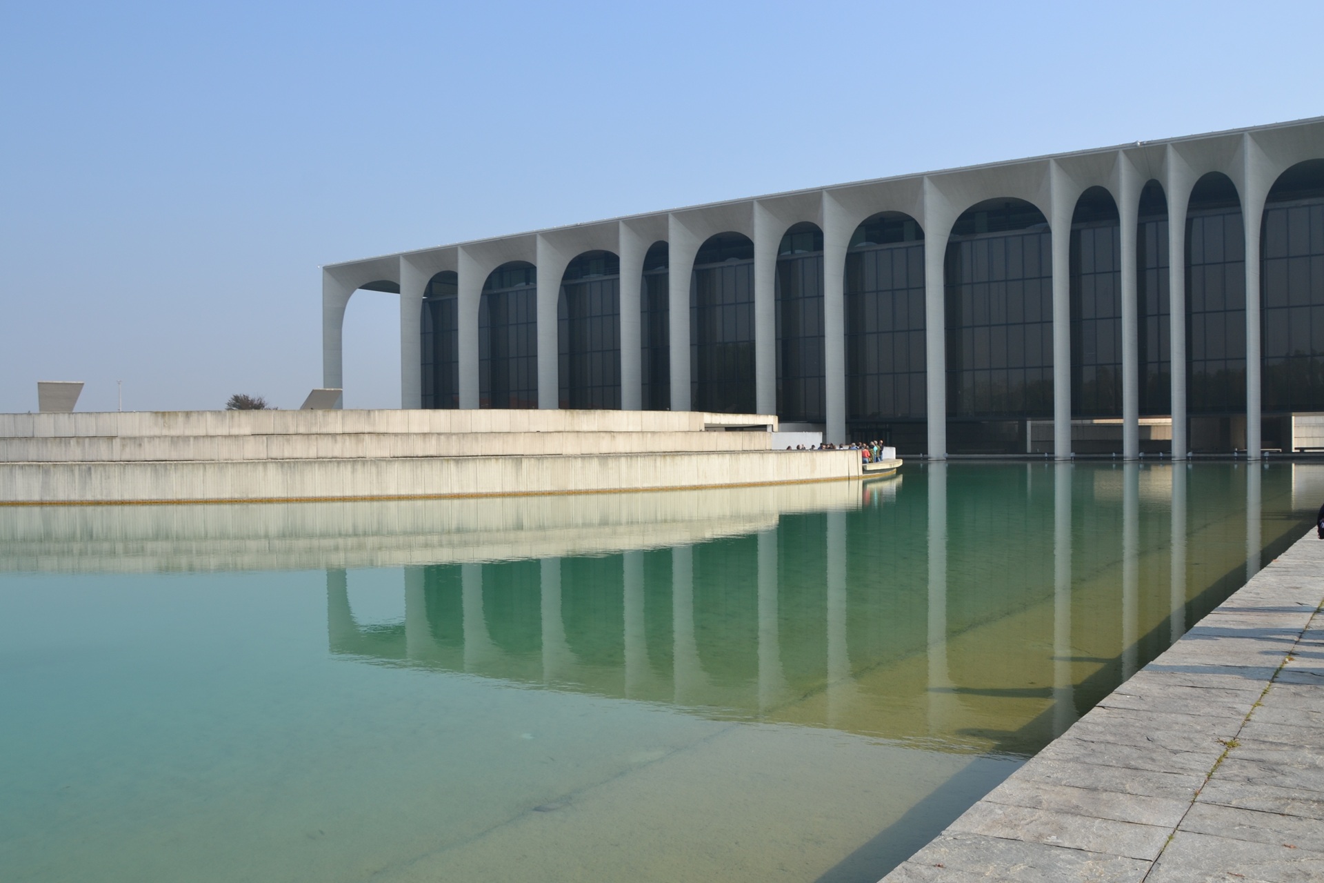 Fotografia do Palácio Mondadori, com arcos altos de concreto alinhados diante de um grande espelho d’água esverdeado; o edifício apresenta fachada envidraçada escura, refletida na superfície calma do lago, sob céu claro e sem nuvens