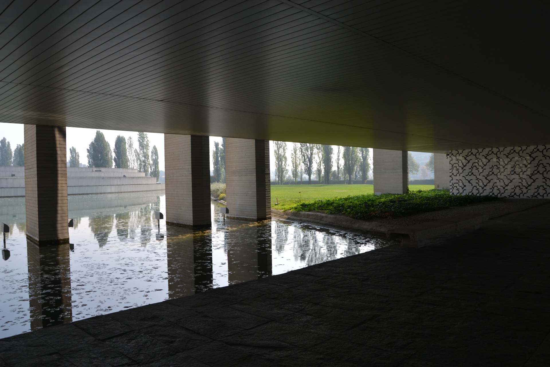 Vista sob a área suspensa do Palácio Mondadori, com pilares refletidos na água rasa; ao fundo, gramado iluminado e uma parede interna revestida pelo padrão característico de Athos Bulcão, composto por formas orgânicas pretas sobre fundo branco