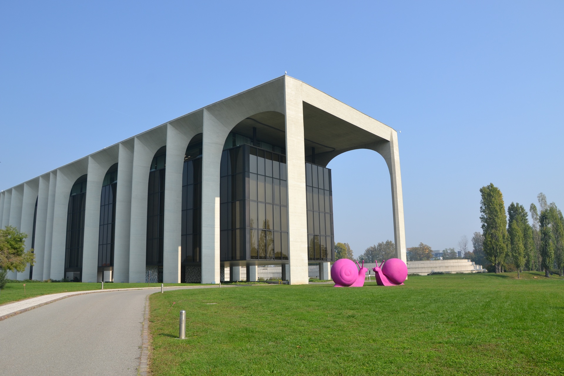 Vista externa do Palácio Mondadori após a revitalização, com grandes arcos de concreto e fachada envidraçada escura; gramado amplo em primeiro plano e céu claro ao fundo