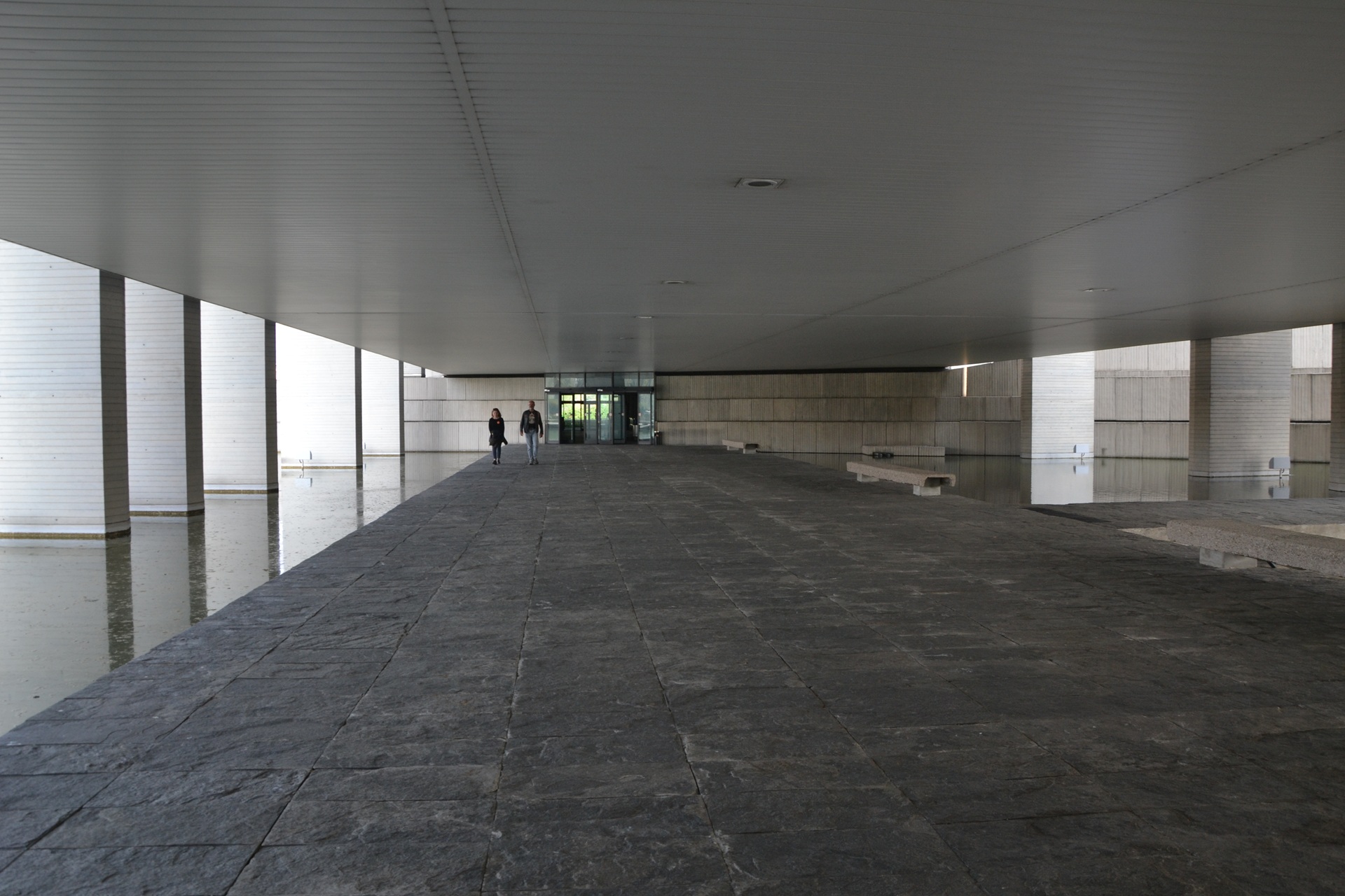 Corredor amplo sob a parte suspensa do Palácio Mondadori, sede da editora; piso de pedra escura, pilares refletidos no espelho d’água e duas pessoas caminhando rumo à entrada envidraçada ao fundo, com luz natural filtrada pelas laterais