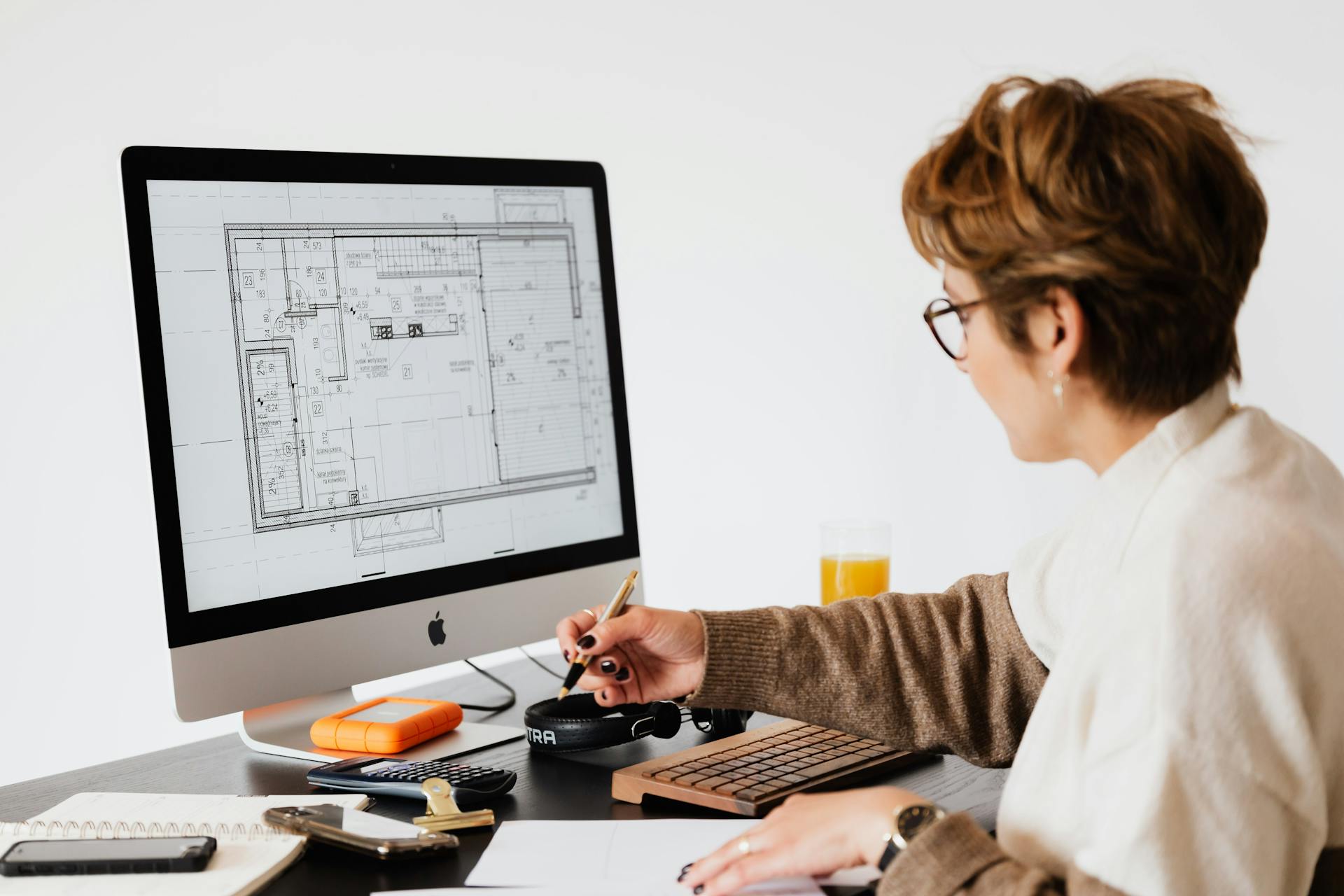 Pessoa sentada à mesa analisando um projeto arquitetônico exibido na tela de um computador grande; ela segura uma caneta e faz anotações, enquanto materiais de trabalho como caderno, calculadora e HD externo estão dispostos ao redor