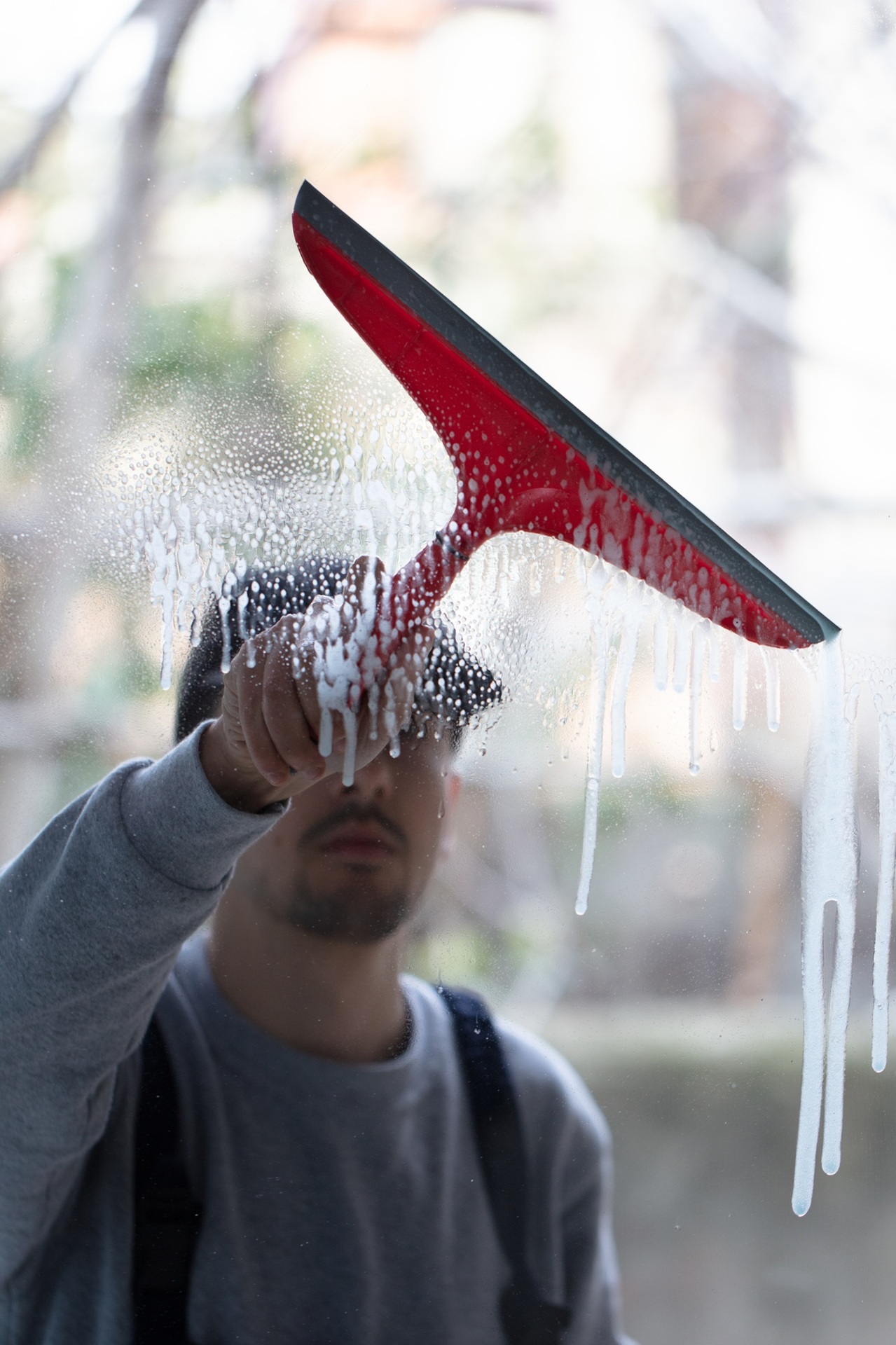 Profissional limpa uma superfície de vidro usando um rodo vermelho com espuma escorrendo; sua figura aparece desfocada atrás do vidro enquanto realiza a limpeza pós-obra