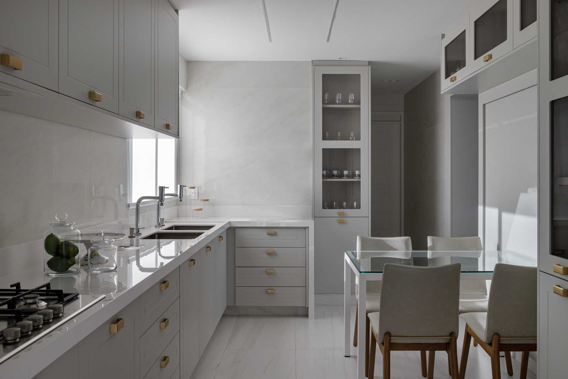 retrospectiva archtrends 2025 Cozinha clara com piso e parede em Onyx Unique, destacando a beleza da pedra calcária; armários cinza com puxadores dourados, bancada branca refletindo luz natural e mesa de vidro com quatro cadeiras claras