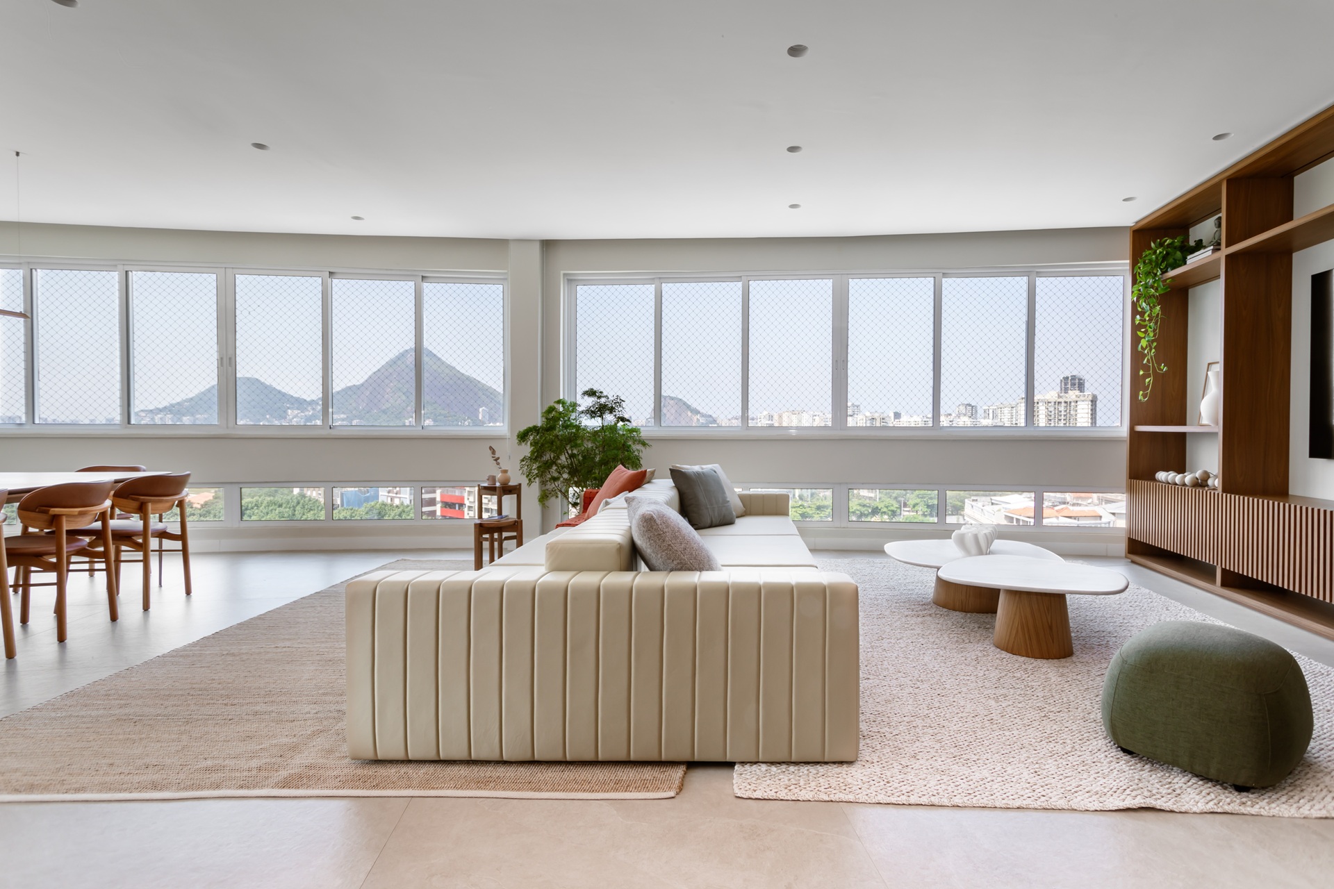retrospectiva archtrends 2025 Sala integrada do Apartamento LO com janelas amplas revelando vista das montanhas, sofá claro de linhas contemporâneas, tapetes texturizados, mesa de centro orgânica e estante amadeirada, destacando atmosfera luminosa, acolhedora e voltada à convivência familiar