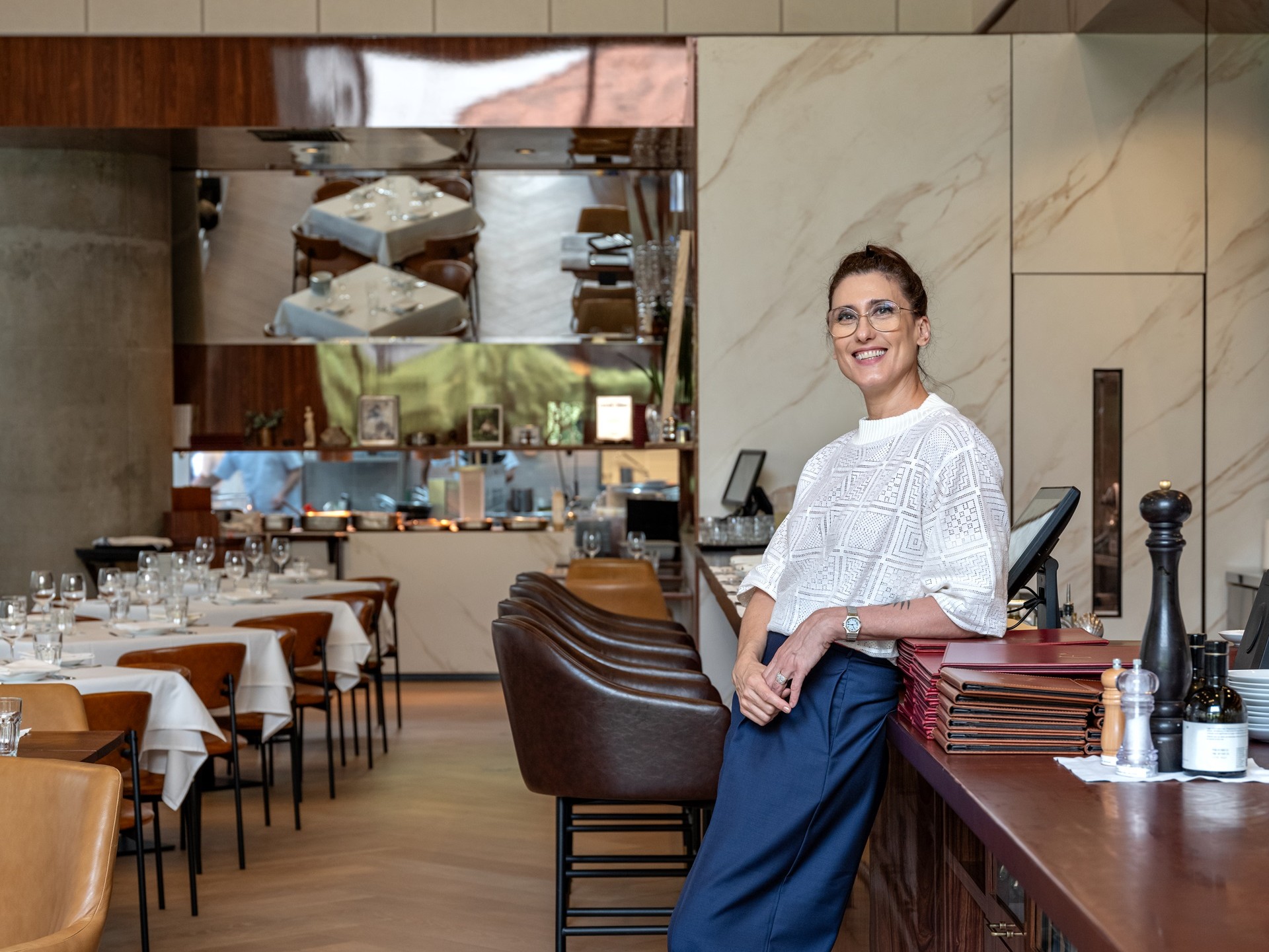 retrospectiva archtrends 2025 Chef Paola Carosella sorrindo ao lado do balcão do restaurante Arturito; interior com mesas postas, cadeiras em couro, painéis em porcelanato Portobello com veios marmorizados e detalhes em madeira, destacando a decoração refinada realizada com produtos da marca