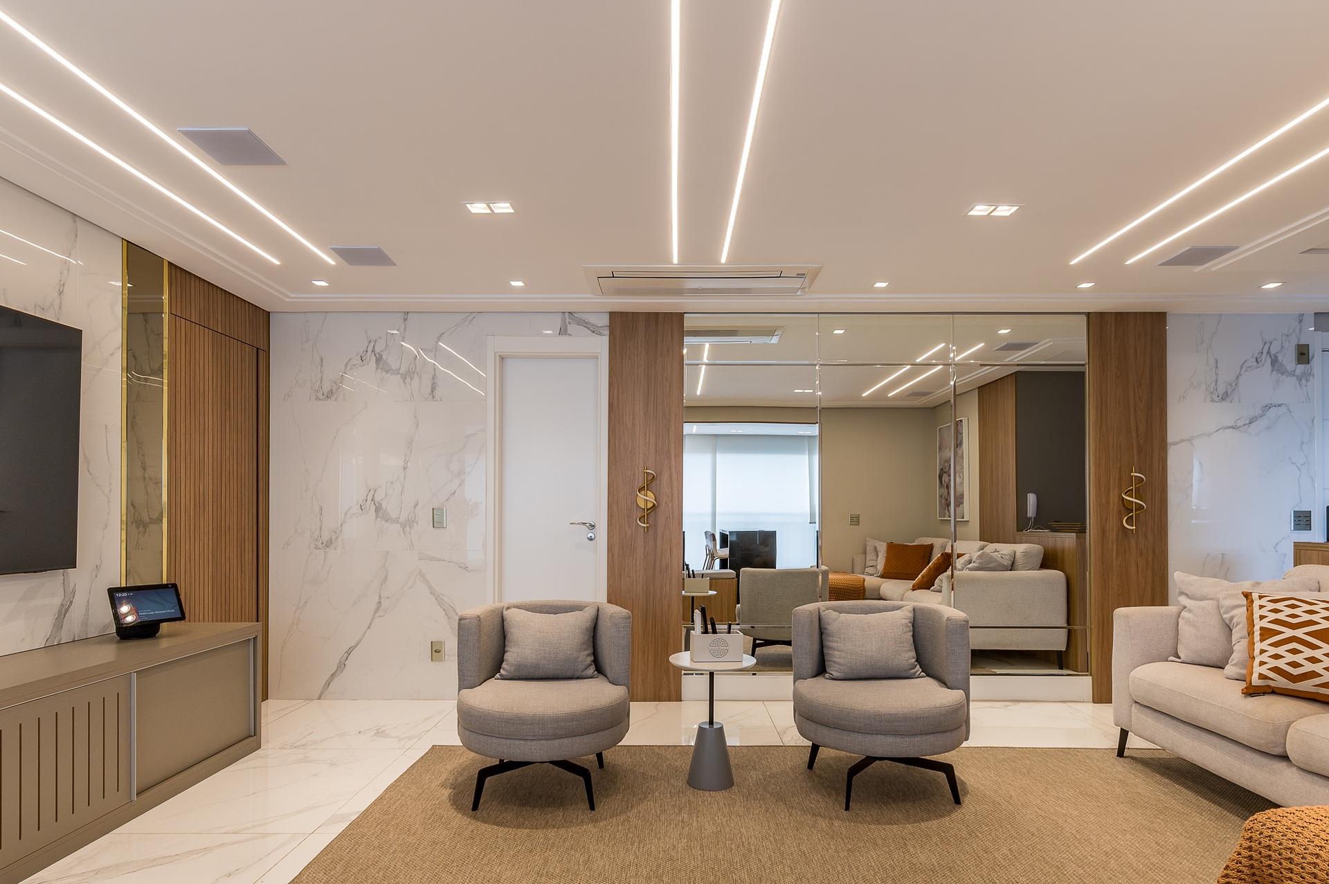 retrospectiva 2025 Sala de estar do Projeto A&P com piso e paredes em revestimento claro, tons dourados nos detalhes, painéis de madeira, poltronas cinza, sofá claro com almofadas e iluminação linear no teto, transmitindo elegância e sofisticação