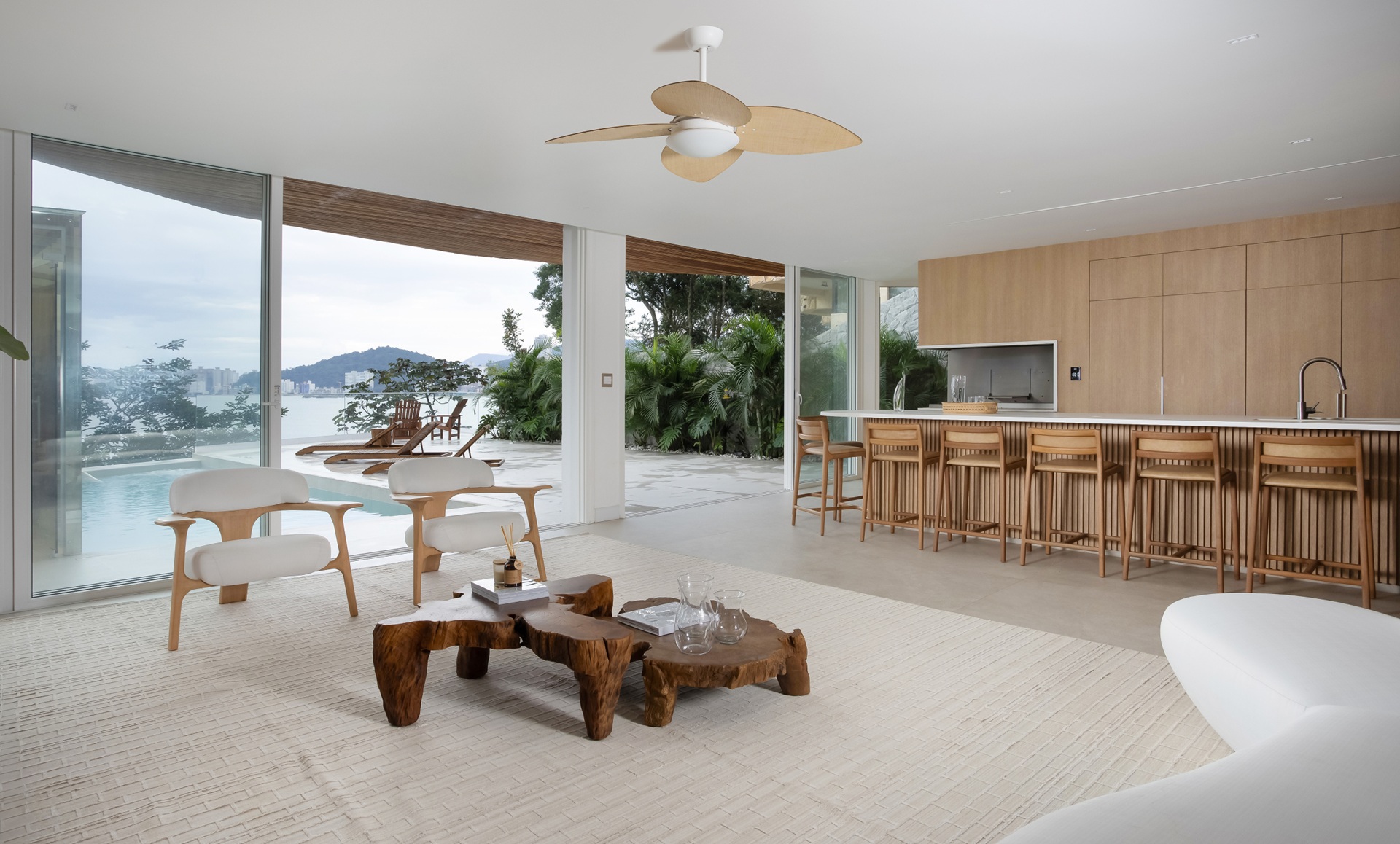 retrospectiva archtrends Living do Projeto Canto do Mar com móveis de design minimalista, mesa de centro em madeira natural, cadeiras claras, cozinha integrada com marcenaria em tom amadeirado, grandes aberturas de vidro para área externa com piscina e vista, destacando amplitude, luz natural e conexão com a natureza