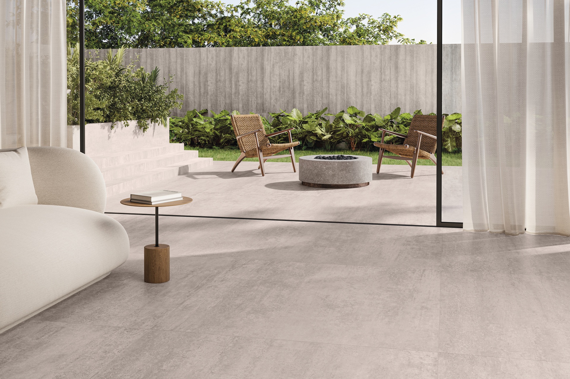 retrospectiva 2025 Ambiente integrado com piso da Linha Síntese em tom claro, sofá curvo creme, mesa lateral de madeira, varanda aberta para área externa com lareira circular, cadeiras naturais e jardim ao fundo, destacando o visual do concreto reinterpretado com suavidade