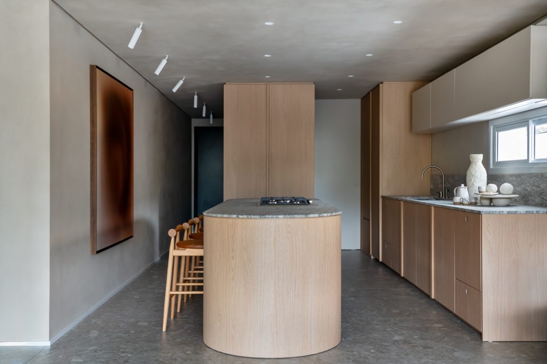 Cozinha com ilha arredondada, mobiliário em madeira clara e piso que reproduz pedra