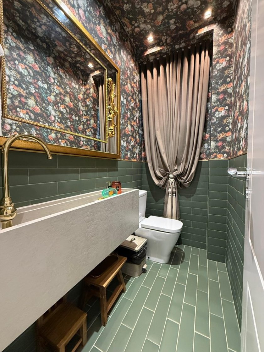 Lavabo com revestimento verde no piso e em meia parede, que também tem papel de parede florido. Metais dourados e uma cortina completam a composição