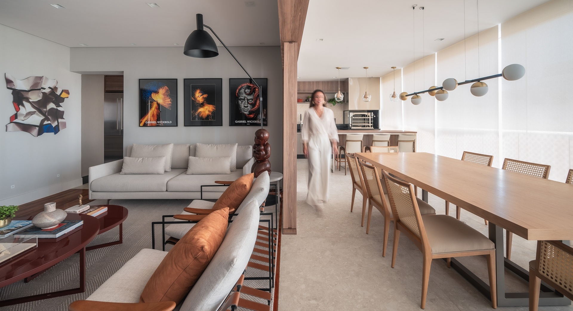 Living integrado à sala de jantar, tudo em tons claros e neutros, com pessoa vestida de branco passando ao lado das cadeiras