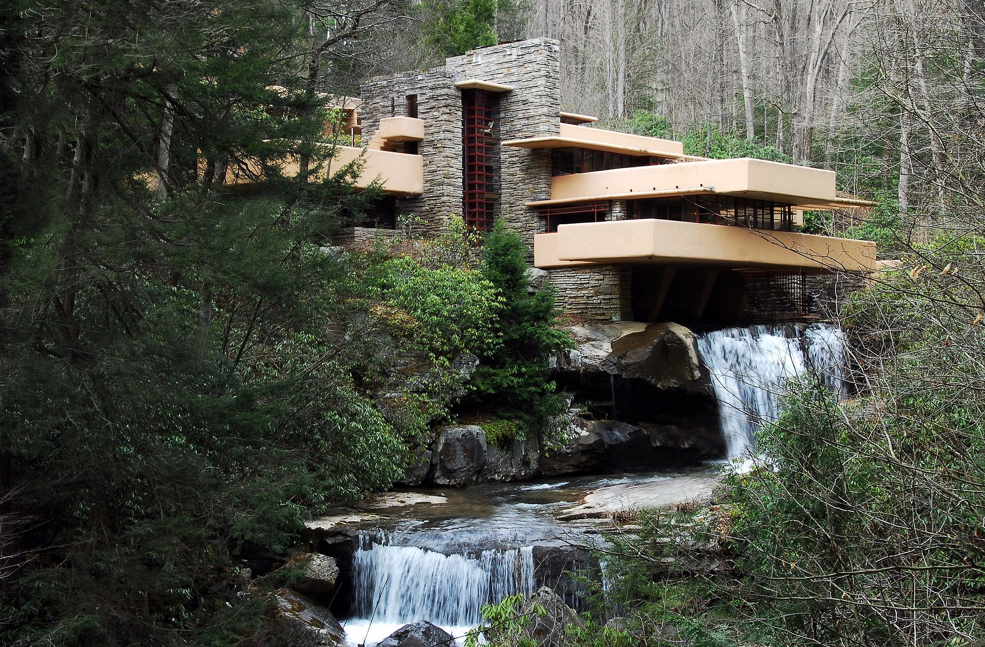 Vista ampla da Fallingwater, residência projetada por Frank Lloyd Wright na Pensilvânia. A construção em concreto e pedra está posicionada sobre uma cachoeira, cercada por vegetação e integrada à paisagem natural
