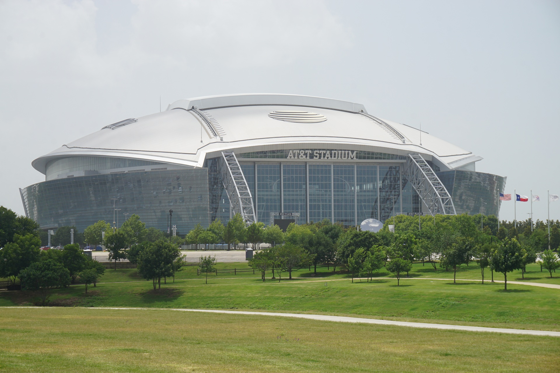 Vista frontal do Dallas Stadium (AT&T Stadium), com cobertura curva branca, fachada envidraçada e área verde em primeiro plano