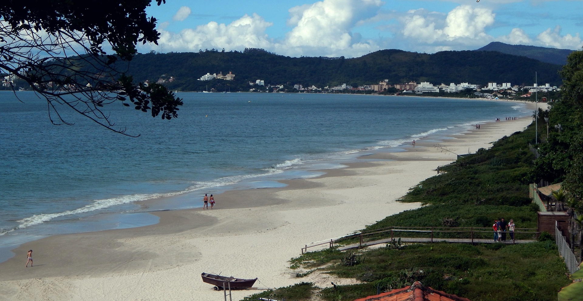Foto da Praia de Jurerê, em Florianópolis, mostrando mar azul-esverdeado, faixa de areia ocupada por banhistas e morros cobertos por vegetação densa ao fundo