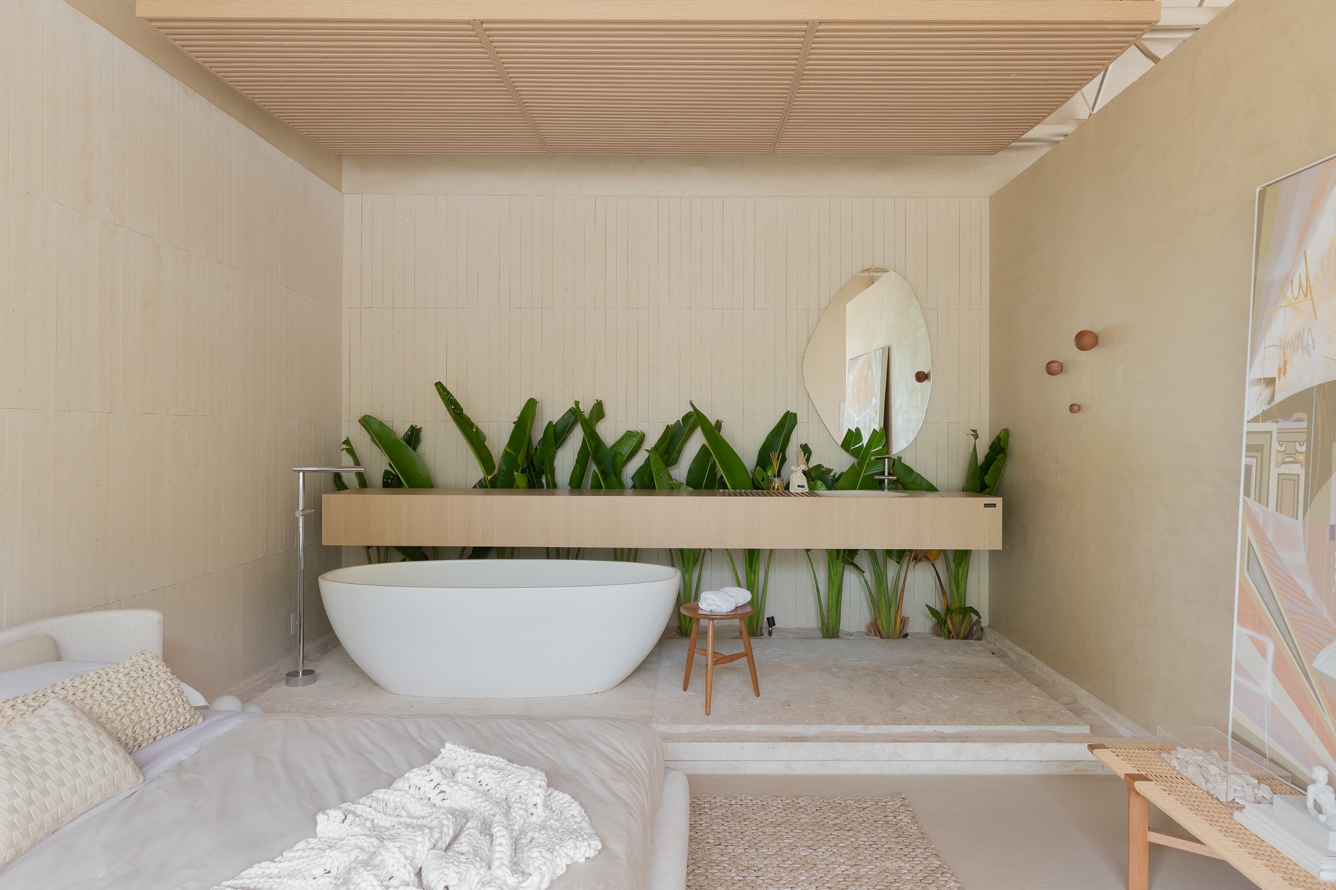 Quarto em tons claros e forro de madeira no teto, com plantas e banheira de imersão ao fundo