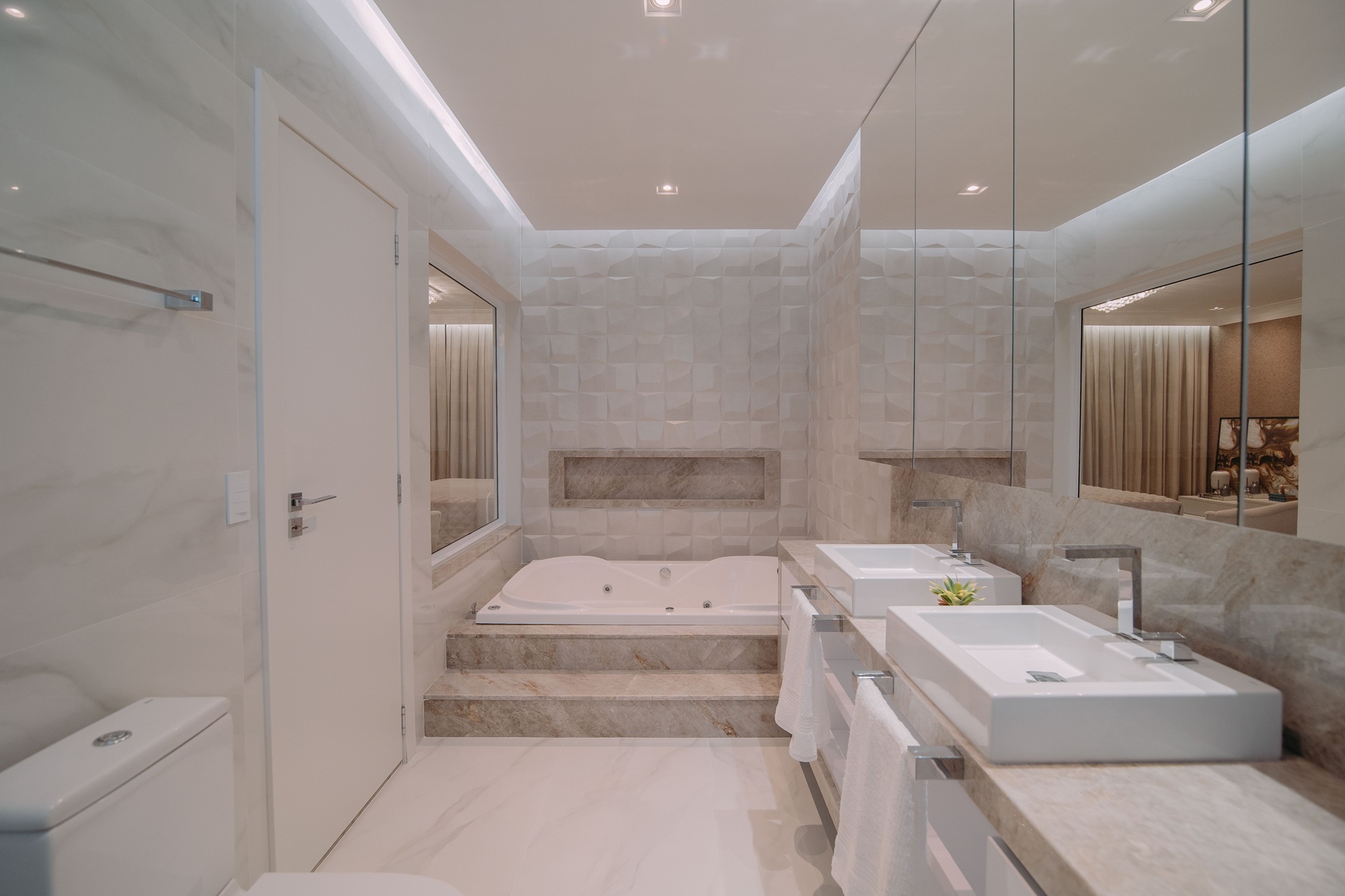 Banheiro com revestimento branco, banheira de hidromassagem com revestimento bege no entorno e parede ao fundo com textura