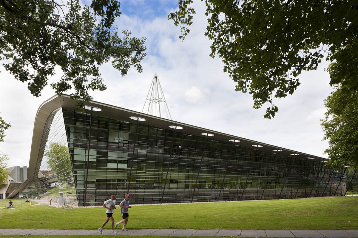 Edifício contemporâneo da Universidade de Tecnologia de Delft, com fachada envidraçada, cobertura curva e estrutura leve. A construção se integra ao gramado e às áreas verdes do campus