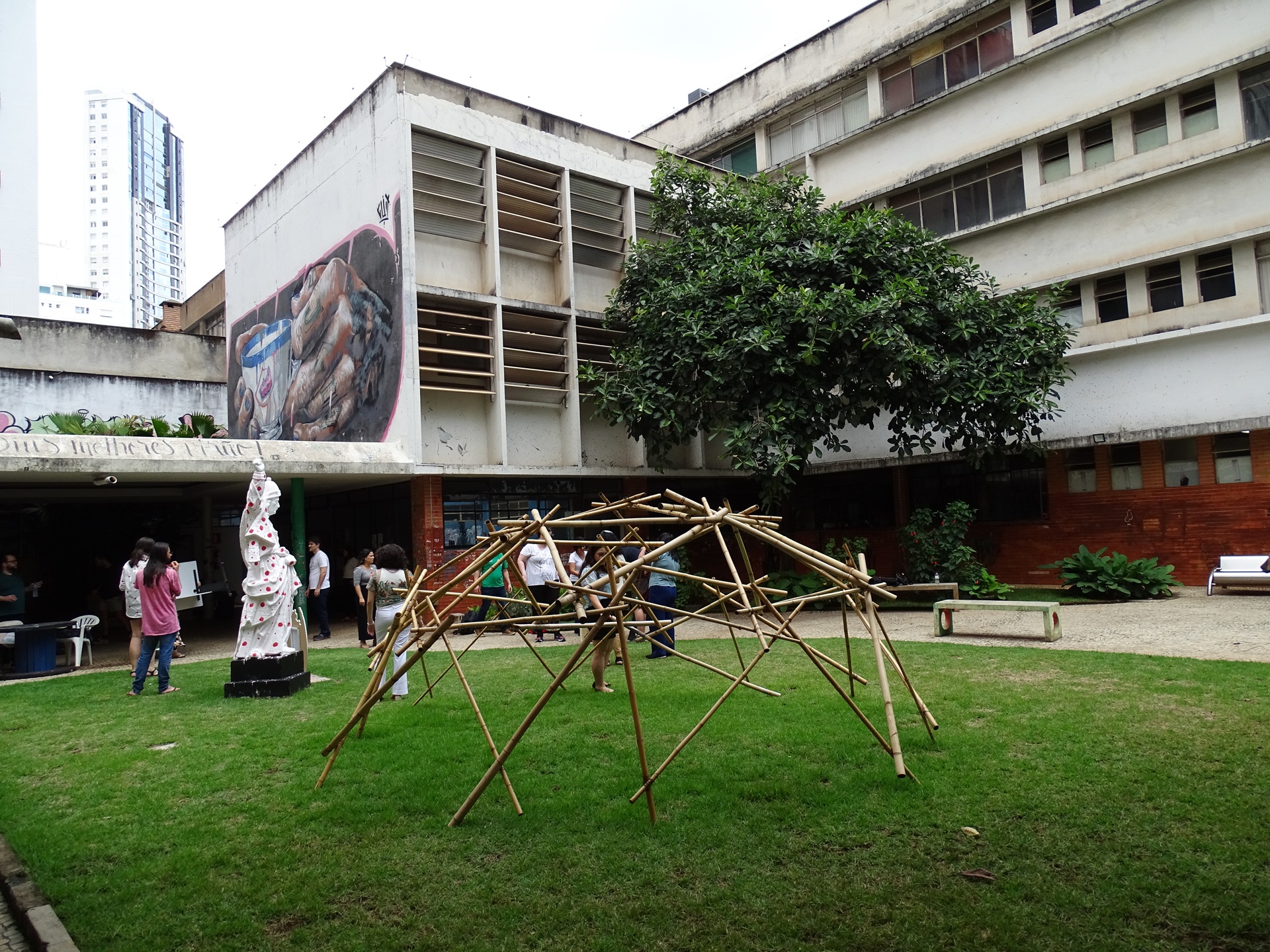 Pátio da Escola de Arquitetura da Universidade Federal de Minas Gerais, com estudantes circulando entre edifícios modernistas. No gramado central, uma instalação estrutural em bambu usada em atividade acadêmica, evidenciando práticas experimentais e o uso do espaço coletivo