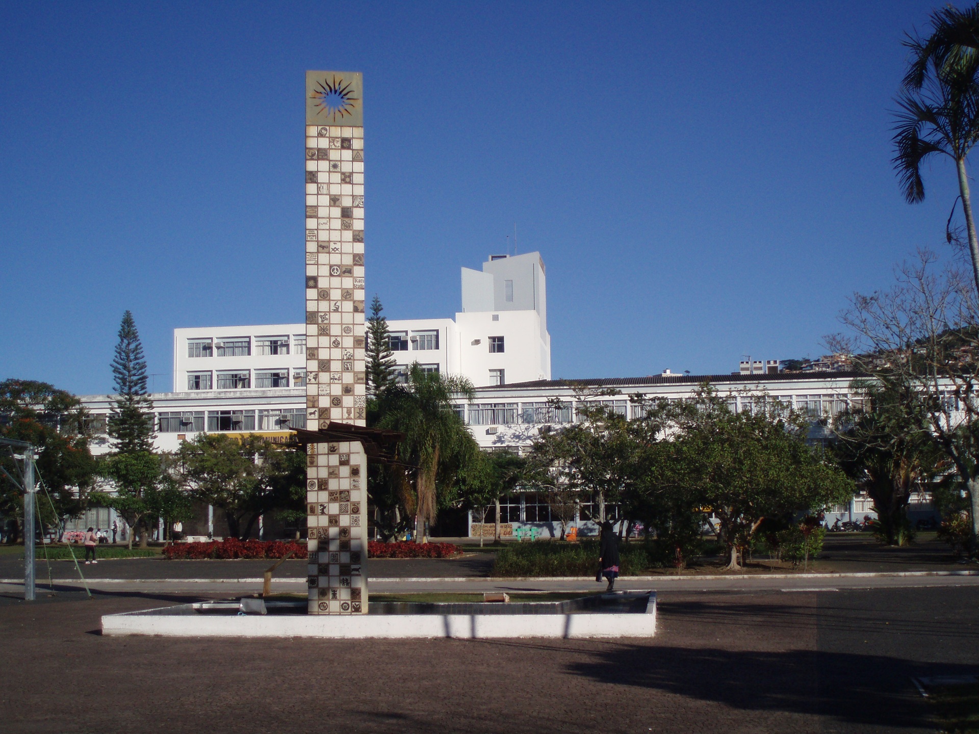 Vista do campus da Universidade Federal de Santa Catarina, com edifícios acadêmicos ao fundo e elemento escultórico vertical em destaque no espaço aberto