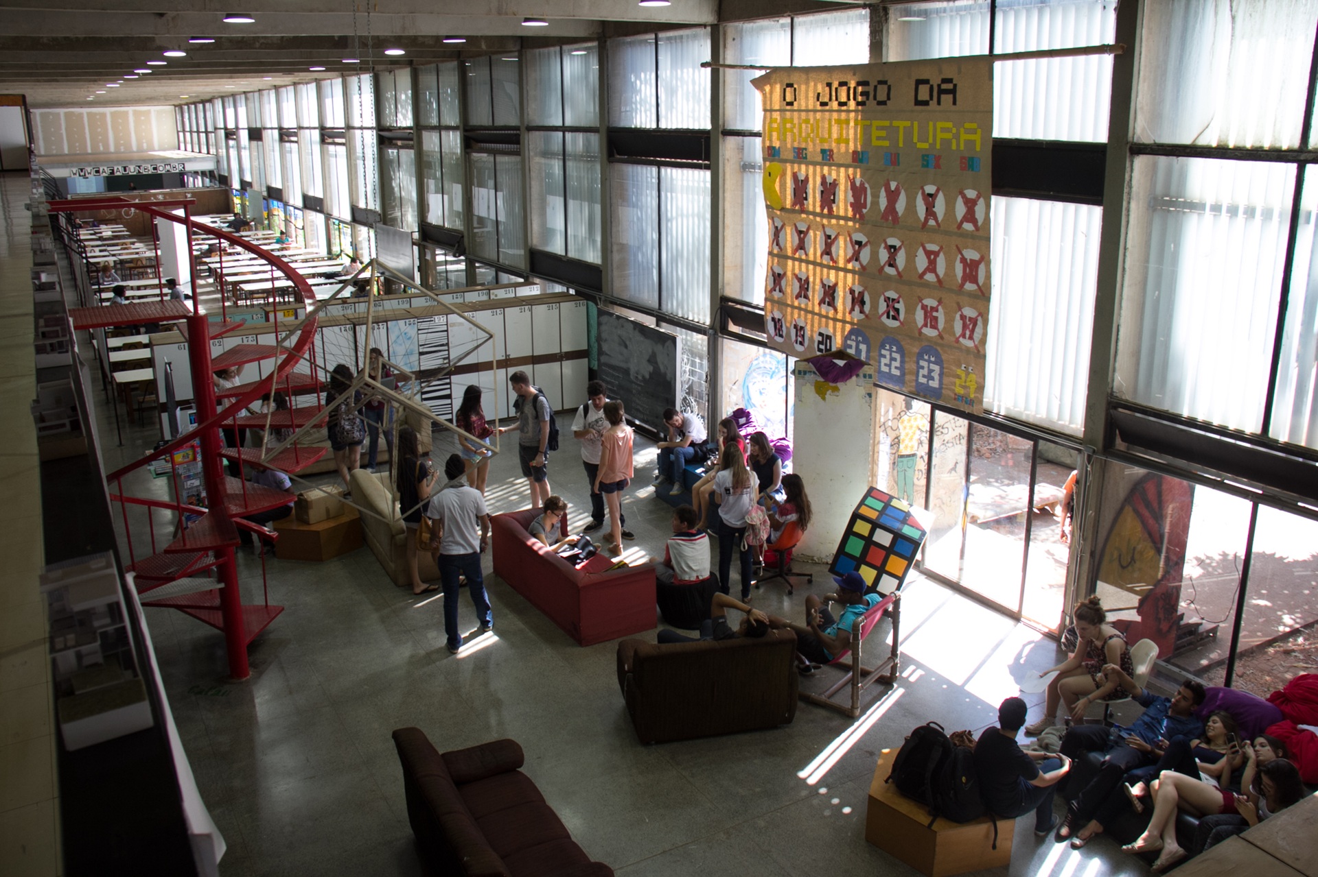 Área interna de uma faculdade de Arquitetura, com pé-direito alto, iluminação natural e estrutura em concreto aparente. Estudantes ocupam sofás e áreas de convivência, interagindo e trabalhando em grupo, enquanto painéis, maquetes e intervenções visuais compõem o ambiente acadêmico e criativo