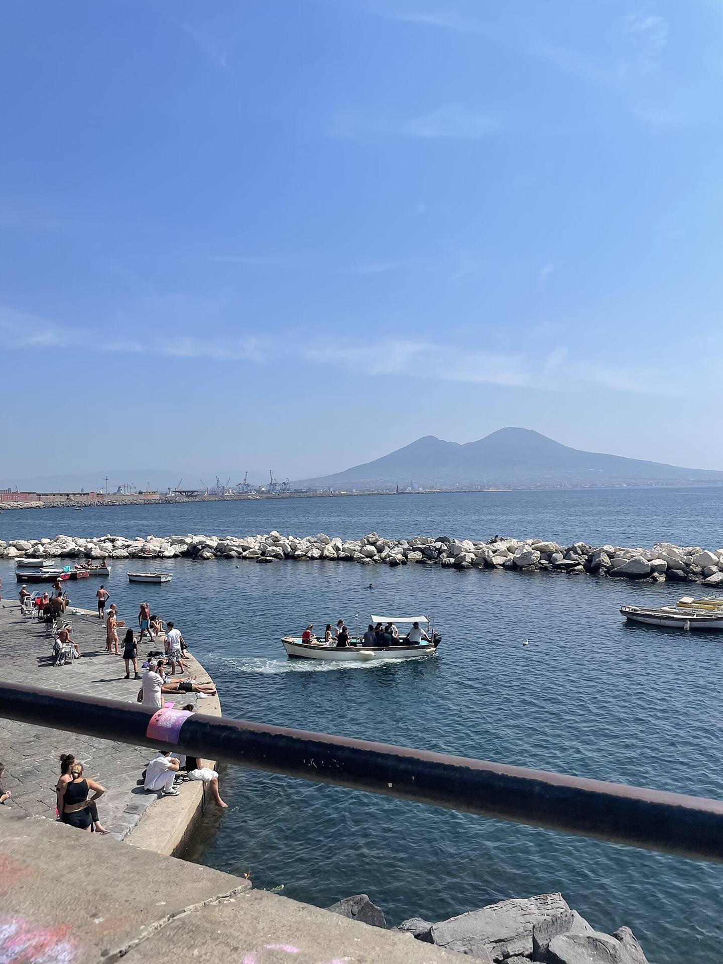 Orla do Lungomare Caracciolo em dia ensolarado, com pessoas sentadas e passeando à beira-mar, pequenos barcos na água e o Vesúvio ao fundo, destacando a paisagem costeira típica de Nápoles