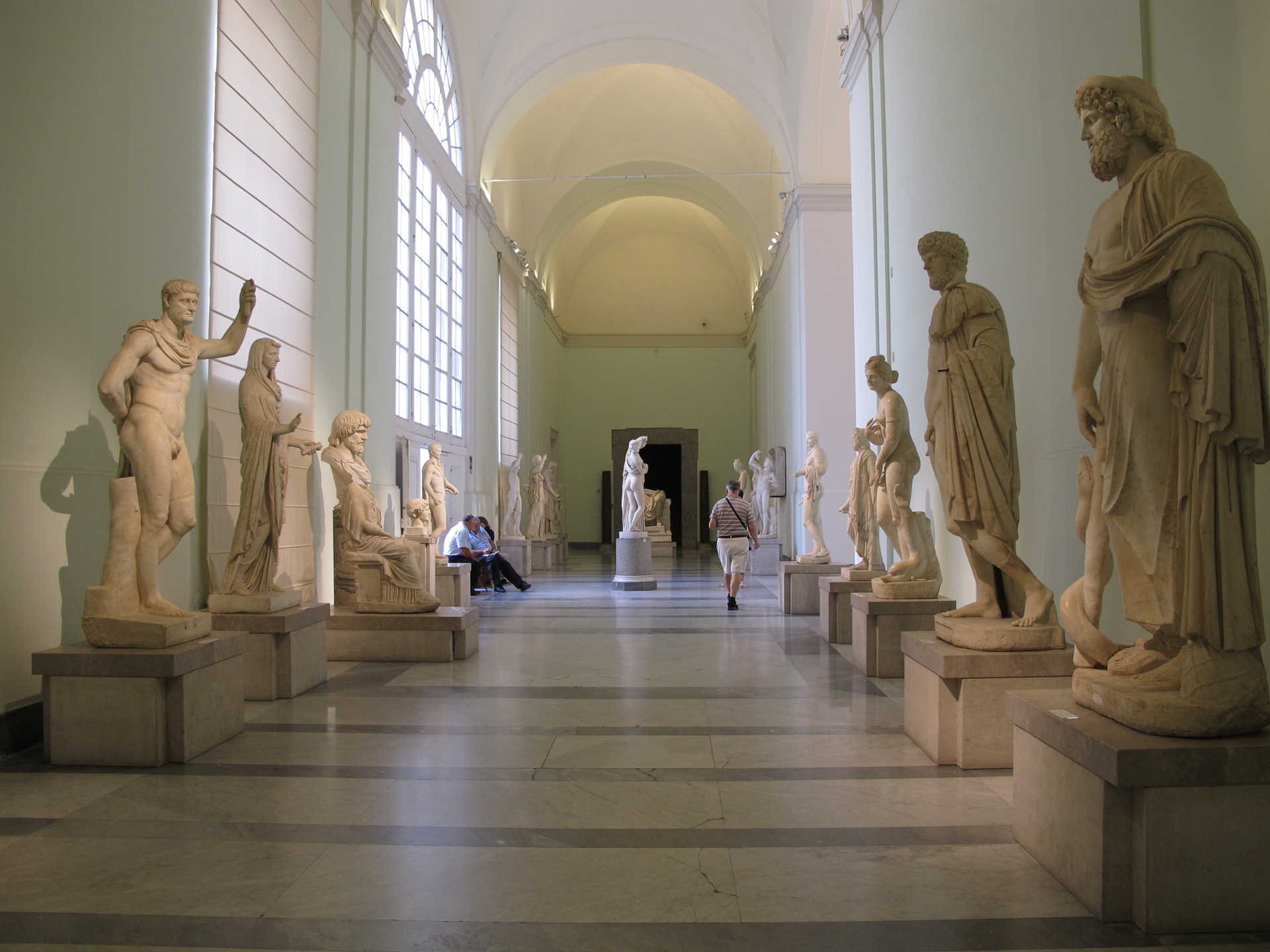 Galeria iluminada do Museu Arqueológico Nacional de Nápoles, com esculturas clássicas alinhadas dos dois lados e visitantes caminhando pelo corredor, destacando a importância histórica e artística do acervo