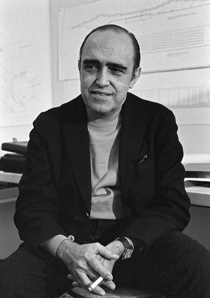 Oscar Niemeyer sentado em seu ambiente de trabalho, segurando um cigarro, com expressão tranquila