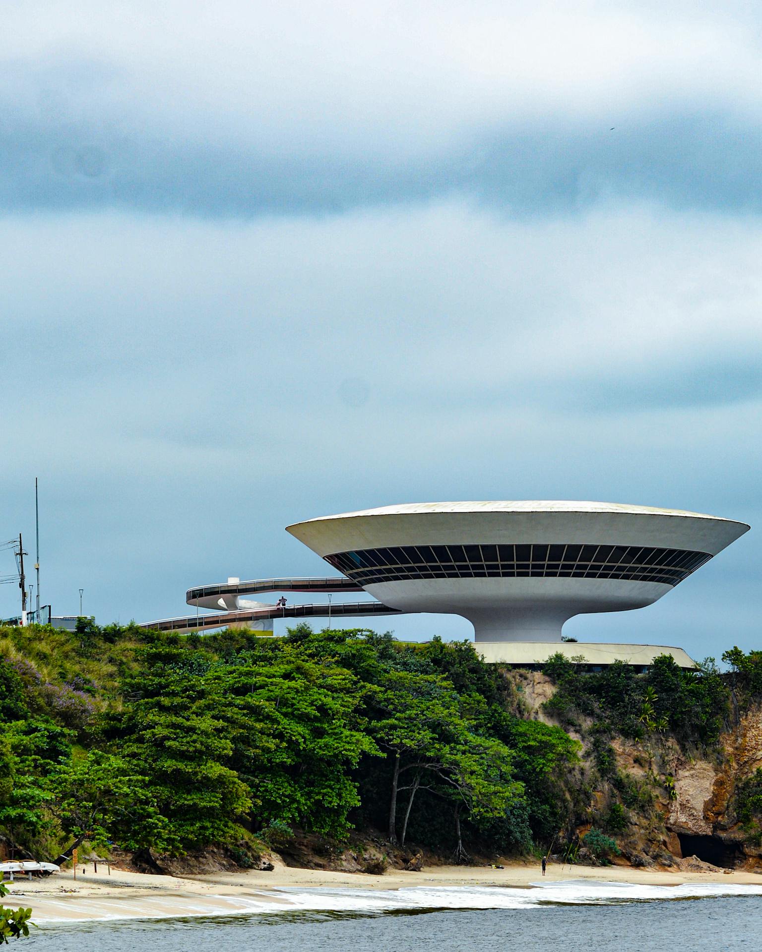 Vista externa do Museu de Arte Contemporânea de Niterói, edifício em forma circular elevado sobre um pilar central, com rampa de acesso sinuosa e implantação sobre falésia à beira da Baía de Guanabara