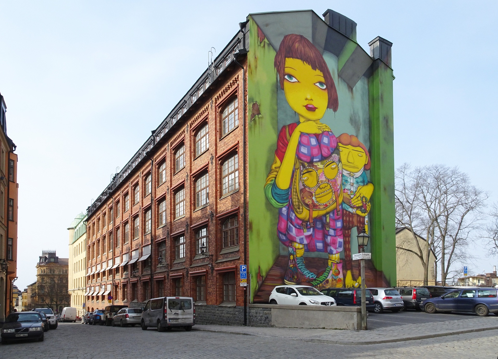osgemeos A lateral de um prédio em Estocolmo exibe um grande mural colorido com personagens amarelos de estilo onírico, incluindo uma figura feminina dominante e outras menores ao redor. Eles usam roupas estampadas em tons vibrantes. A cena aparece sobre uma rua de paralelepípedos com carros estacionados à frente