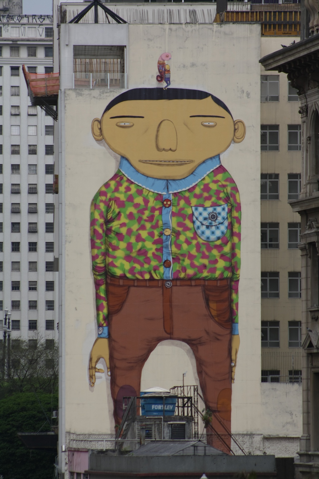 os gemeos A imagem mostra um grande mural em um prédio no centro da cidade, retratando um personagem estilizado de pele amarela e expressão calma. Ele veste camisa estampada em cores vivas, calça marrom e tem um pequeno boneco equilibrado sobre sua cabeça. O mural ocupa toda a lateral do edifício, cercado por outros prédios altos