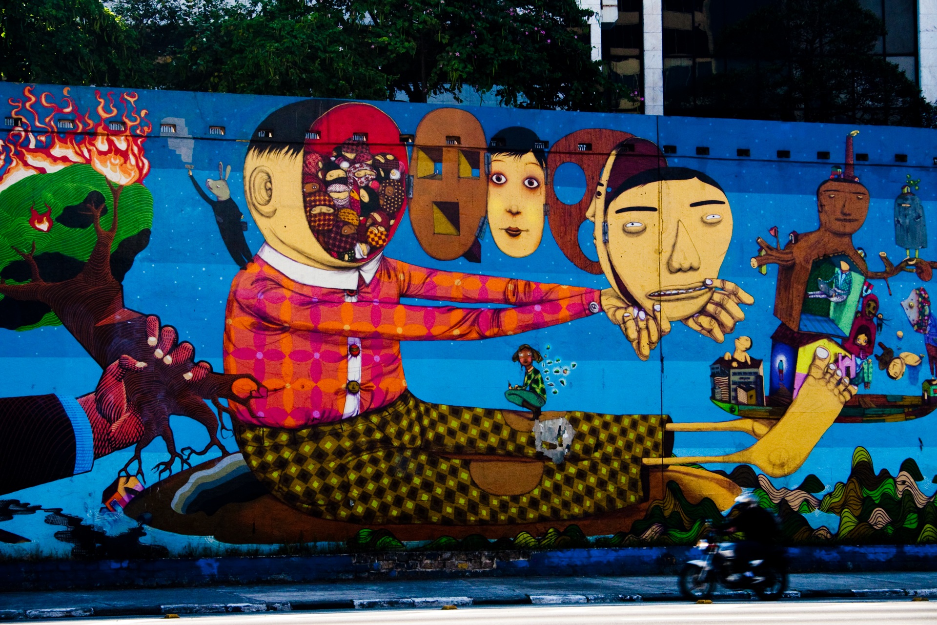 osgemeos O mural mostra uma grande figura estilizada com o rosto dividido entre uma metade aberta, cheia de pequenos objetos coloridos, e outra metade ausente. Ela segura uma máscara humana, enquanto rostos ovais aparecem alinhados ao fundo. À esquerda, há uma árvore em chamas; à direita, personagens e casas surgem em estilo fantástico