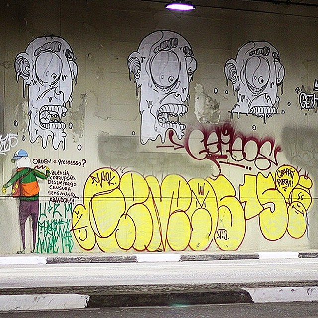 osgemeos A imagem mostra um mural em um túnel com várias intervenções: à esquerda, um personagem colorido escreve palavras como “violência”, “corrupção” e “desemprego”, acompanhadas da pergunta “Ordem e Progresso?”. Acima dele, há desenhos repetidos de rostos grandes em estilo expressivo, e a parte inferior do muro está coberta por tags e grafites amarelos e vermelhos