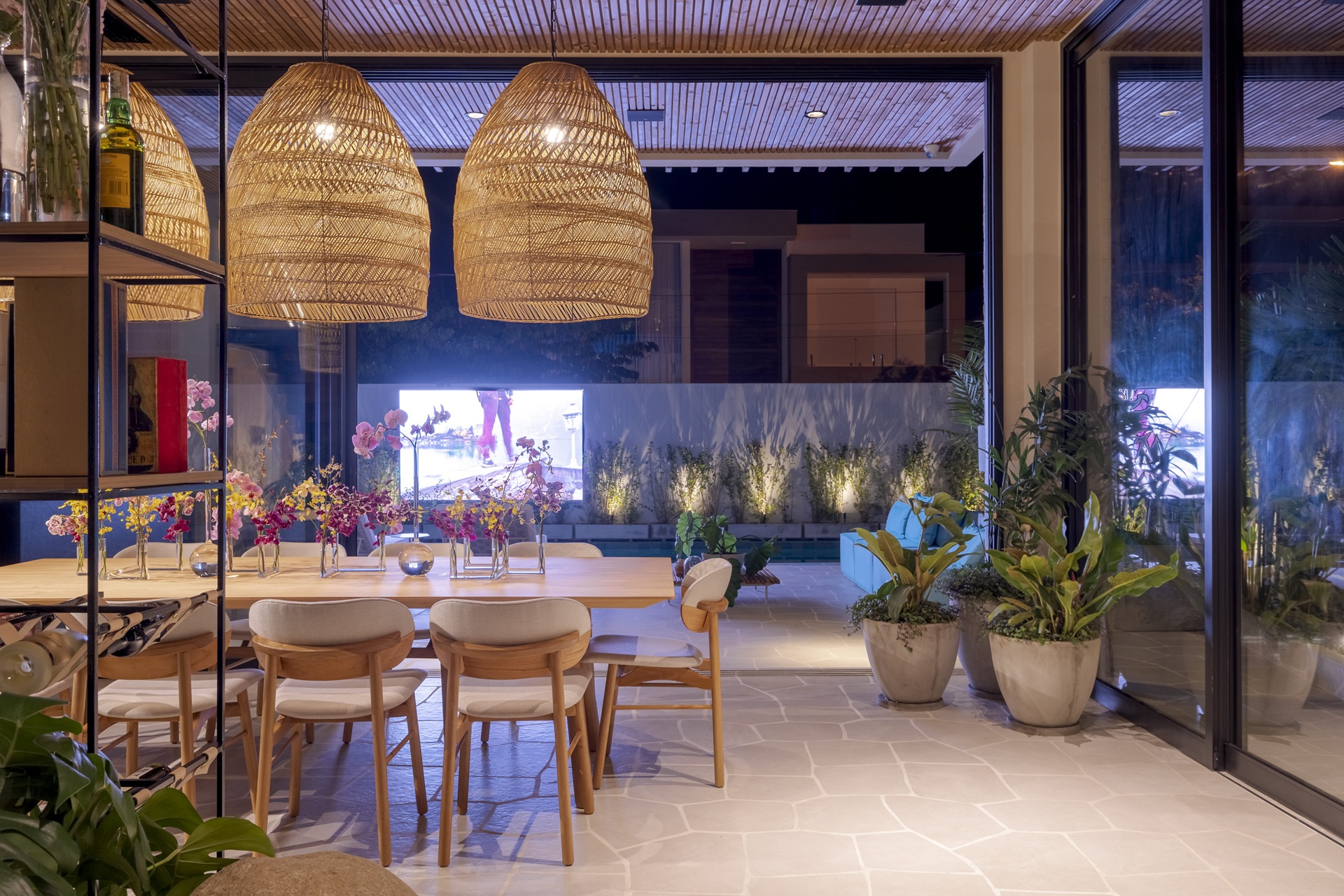 Área externa coberta integrada ao ambiente interno, com mesa de jantar em madeira, cadeiras estofadas e forte presença de vegetação. Pendentes de palha trançada suspensos sobre a mesa garantem iluminação suave e contribuem para uma atmosfera leve e convidativa. O espaço combina materiais naturais, transparências e paisagismo, ideal para momentos de convivência ao ar livre com conforto visual