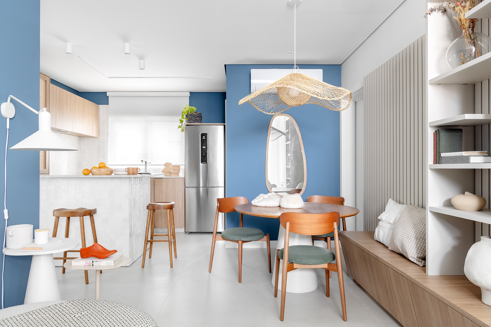 Ambiente integrado de sala de jantar e cozinha com paleta clara e pontos de azul nas paredes. O pendente de palha com desenho orgânico se destaca sobre a mesa redonda, harmonizando com cadeiras em madeira, estofados em tons neutros, marcenaria clara e objetos cerâmicos