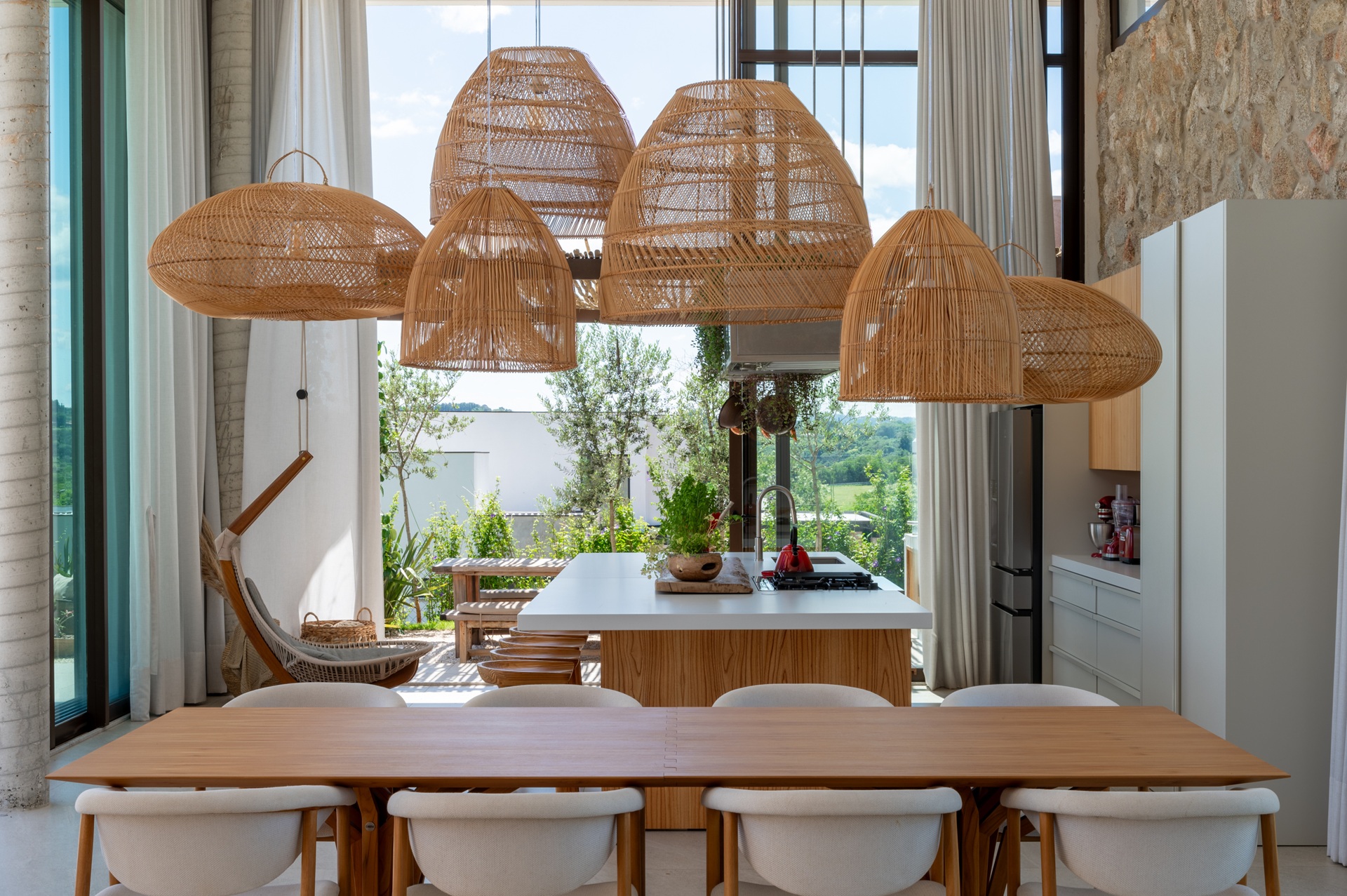 Ambiente integrado de jantar e cozinha com pé-direito alto e ampla entrada de luz natural. Conjunto de pendentes de palha em diferentes formatos, volumes e tramas suspensos sobre a mesa e a ilha criam uma composição dinâmica e equilibrada