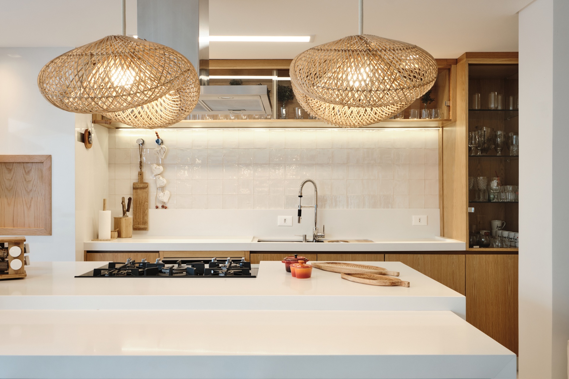 Cozinha contemporânea com ilha central e paleta clara, marcada por superfícies brancas e armários em madeira natural. Dois pendentes de palha trançada suspensos sobre a bancada funcionam como ponto focal e fonte de iluminação suave. A composição equilibra funcionalidade e estética, valorizando a integração entre materiais naturais e soluções atuais de projeto