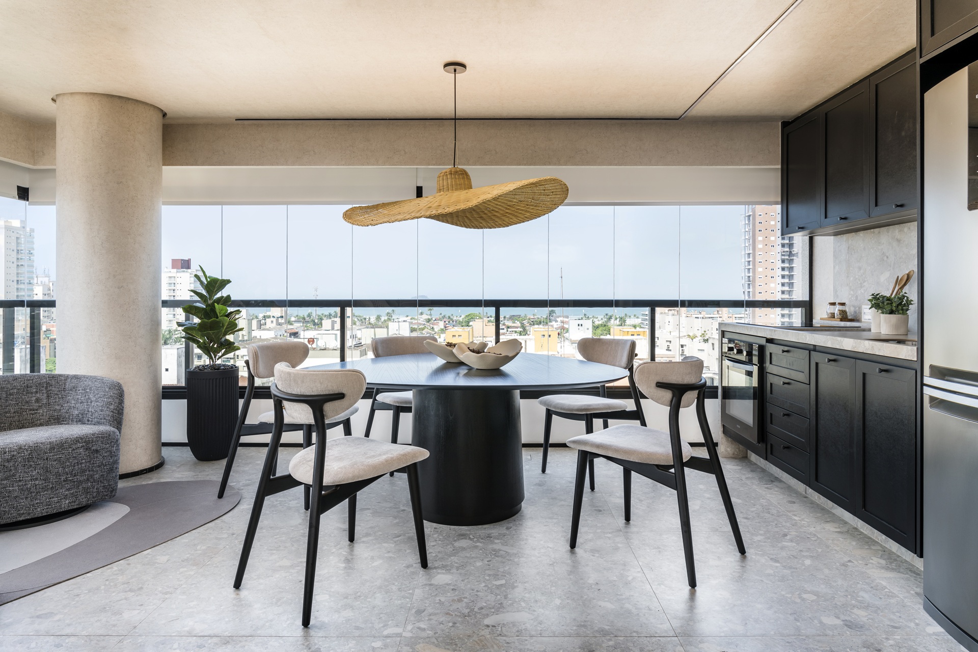 Área integrada de jantar e cozinha com estética contemporânea e vista urbana ao fundo. A mesa central de base escura recebe um pendente de palha de desenho orgânico e alongado, evidenciando a versatilidade da peça em composições sofisticadas. Cadeiras estofadas em tons neutros dialogam com a marcenaria escura, o piso claro e a vegetação pontual, criando um ambiente equilibrado, funcional e visualmente leve
