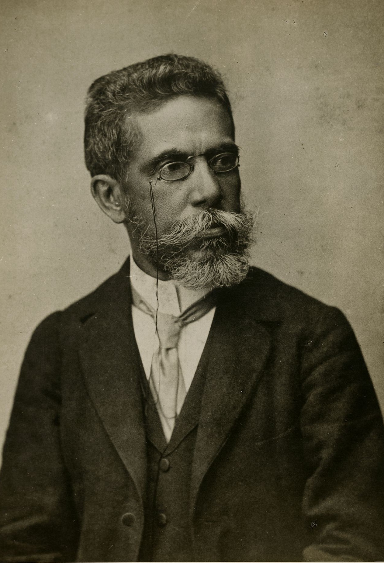 Retrato em preto e branco de Machado de Assis aos 57 anos, feito em 1896. O escritor aparece de terno escuro, colete e gravata clara, usando pince-nez (óculos com suporte nasal). Seu cabelo está curto e grisalho, assim como a barba espessa e bem aparada. Ele olha levemente para o lado, com expressão calma e pensativa