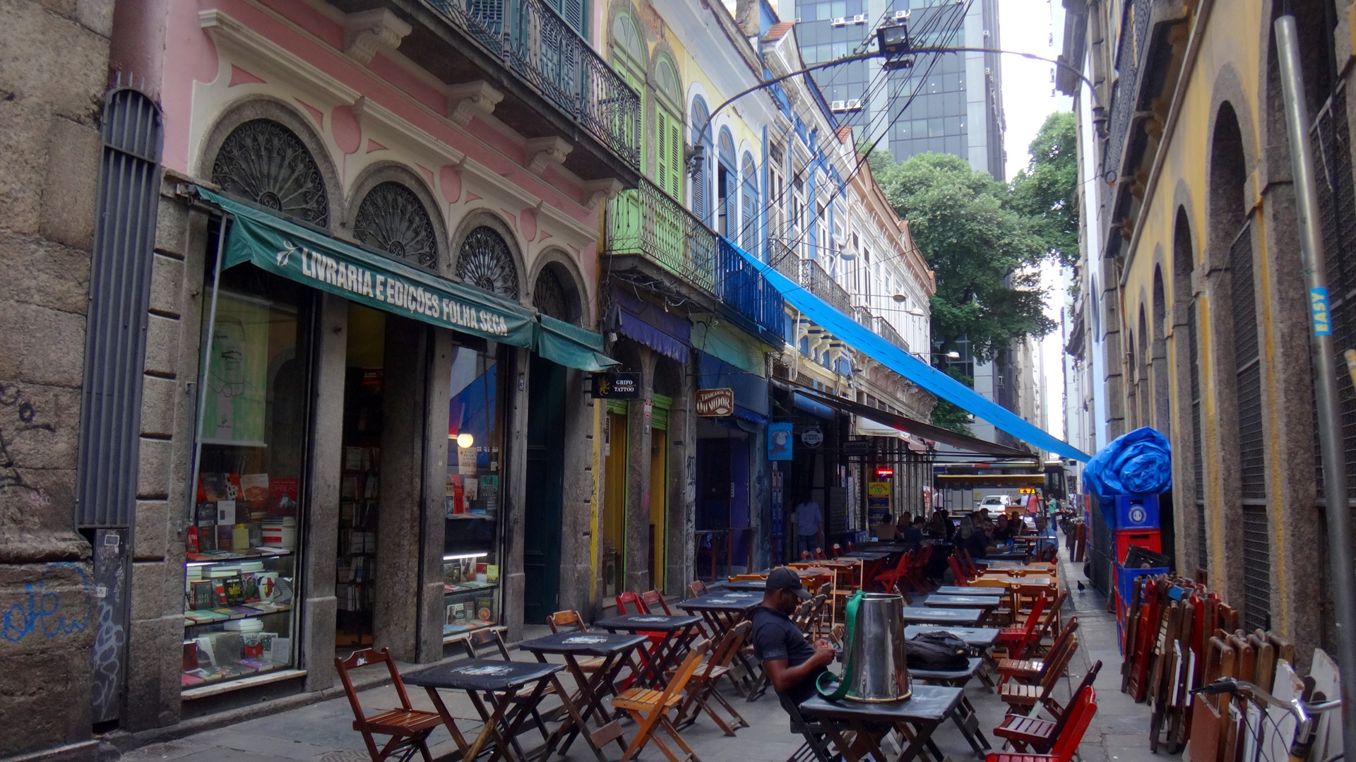 Vista da Rua do Ouvidor, no centro do Rio de Janeiro, com mesas de bares ao ar livre, fachadas coloridas e varandas de ferro trabalhado. À esquerda, uma livraria ocupa o térreo de um prédio histórico com janelas arqueadas