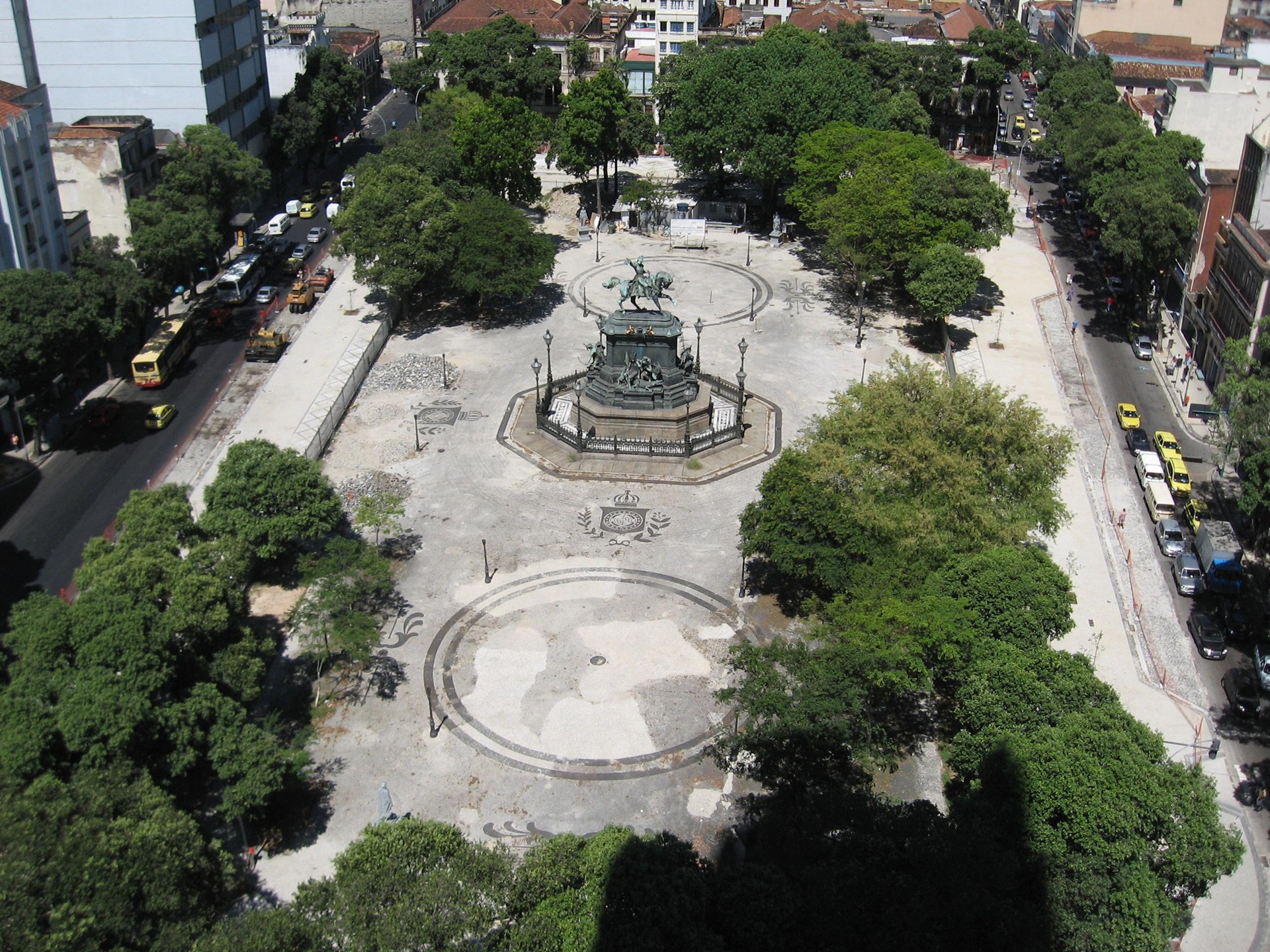 Vista aérea da Praça Tiradentes, no centro do Rio de Janeiro, com árvores ao redor e a estátua equestre de Dom Pedro I no centro, cercada por calçamento decorativo e edifícios históricos nas ruas adjacentes