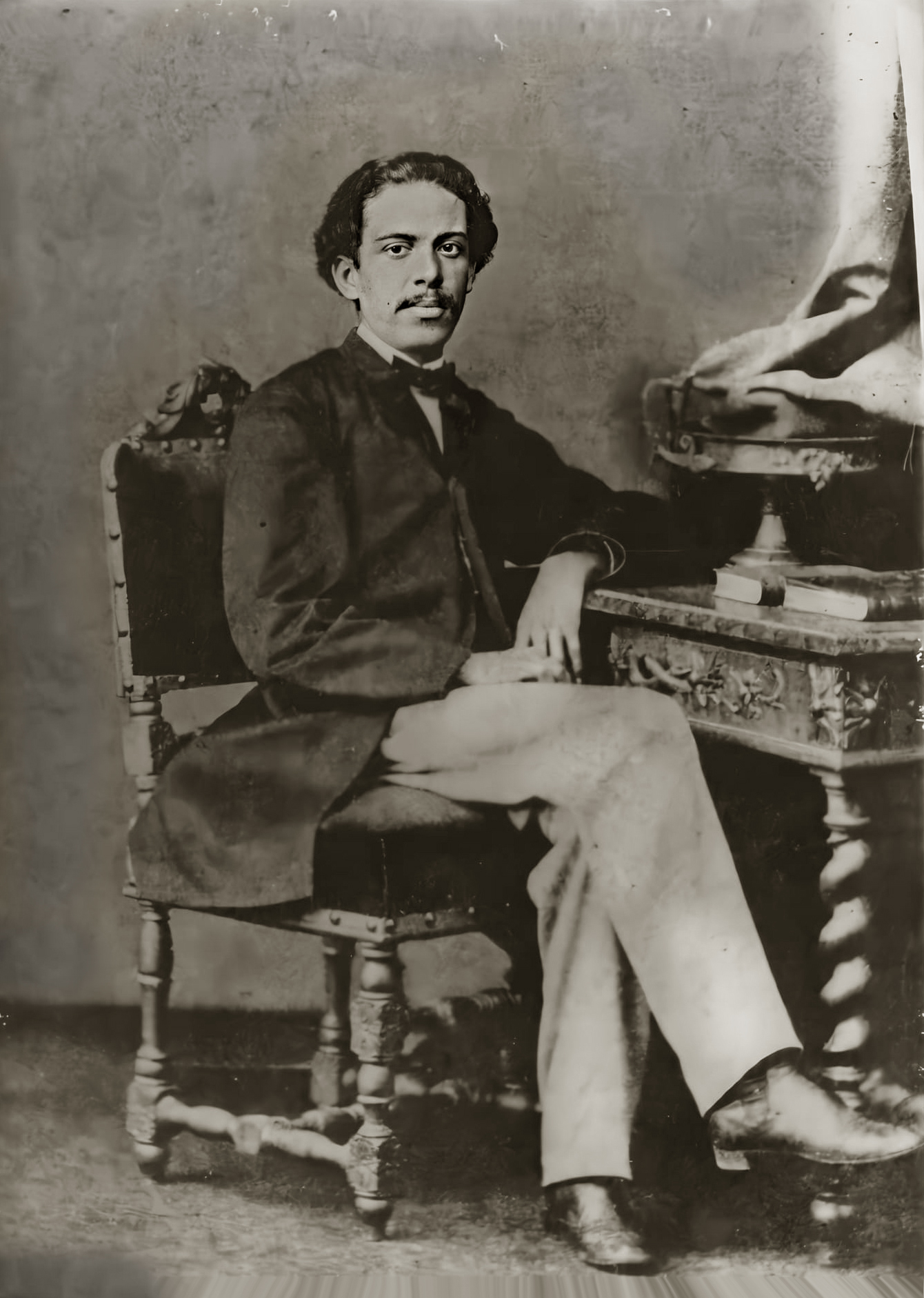 Retrato em preto e branco de Machado de Assis aos 25 anos, feito em 1864. Ele está sentado em uma cadeira de madeira entalhada, usando paletó escuro, calça clara e gravata. O escritor posa ao lado de uma mesa ornamentada, apoiando o braço direito sobre o tampo, em uma postura formal e serena