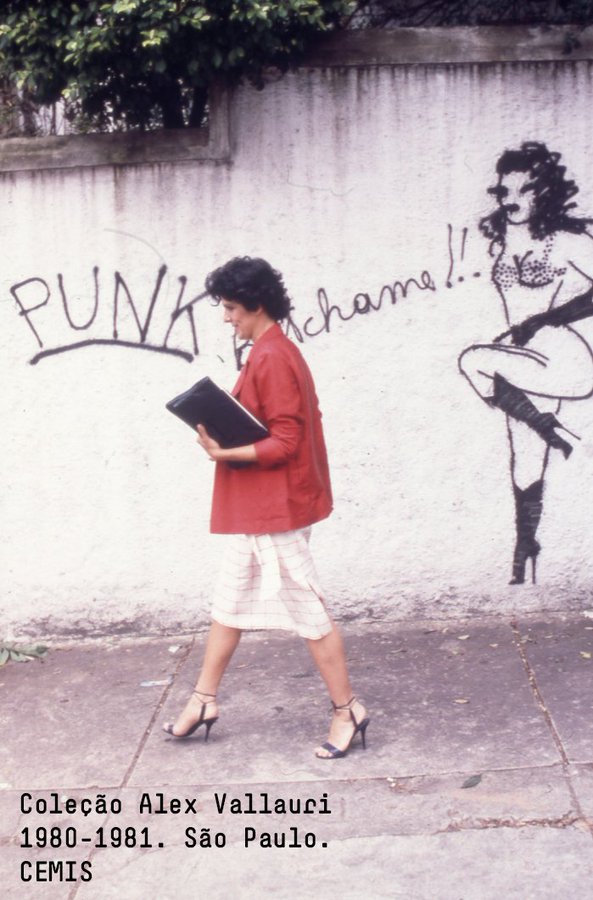 street art Fotografia de uma mulher caminhando diante de um muro com grafite de Alex Vallauri, feito entre 1980 e 1981 em São Paulo. A pintura mostra uma figura feminina provocante ao lado da palavra “PUNK” e da frase “me chame!!”
