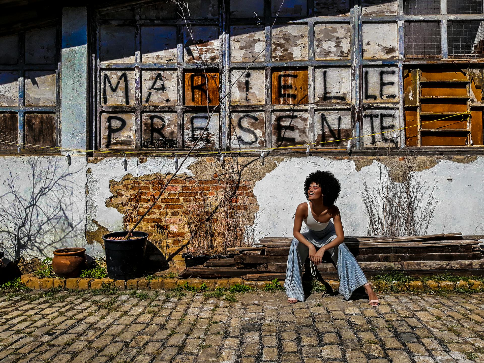 street art Mulher negra sentada e sorrindo diante de um muro com a frase “Marielle Presente” pintada em letras pretas sobre janelas antigas